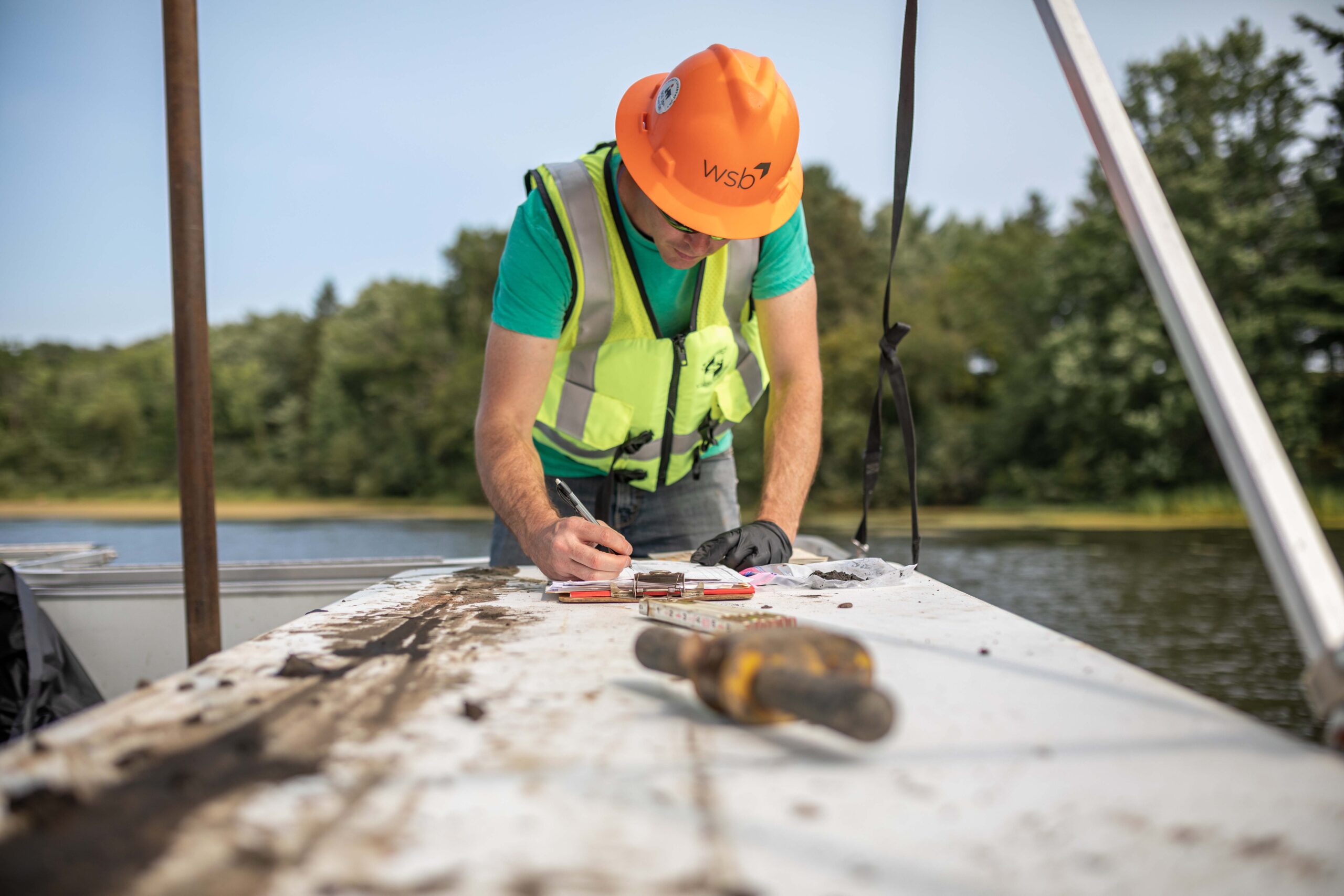 Engineer wearing safety gear writing on a piece of paper on a boat on a lake.