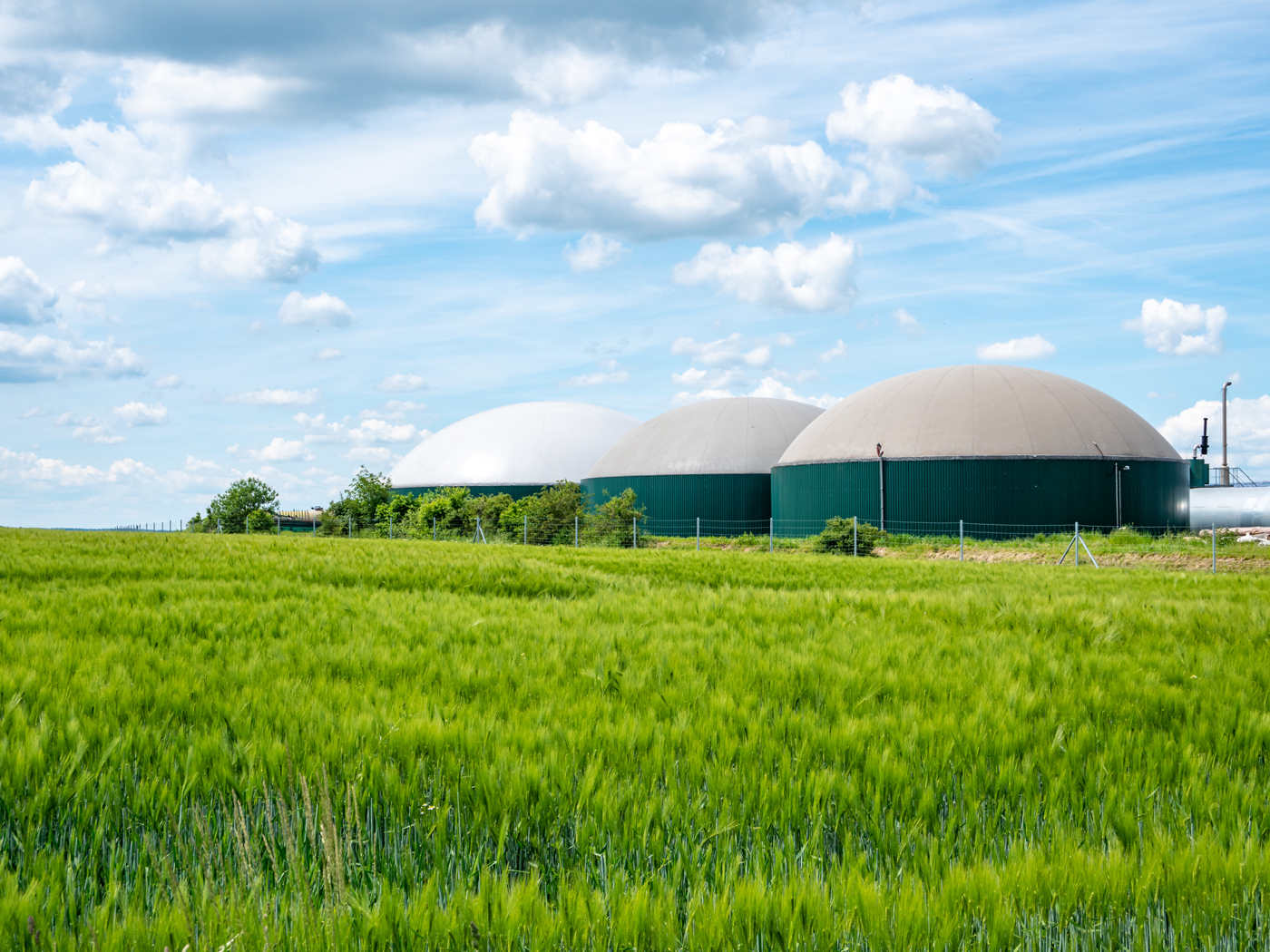 Three large dome-shaped biogas structures in a rural landscape with a green field and a partly cloudy sky.