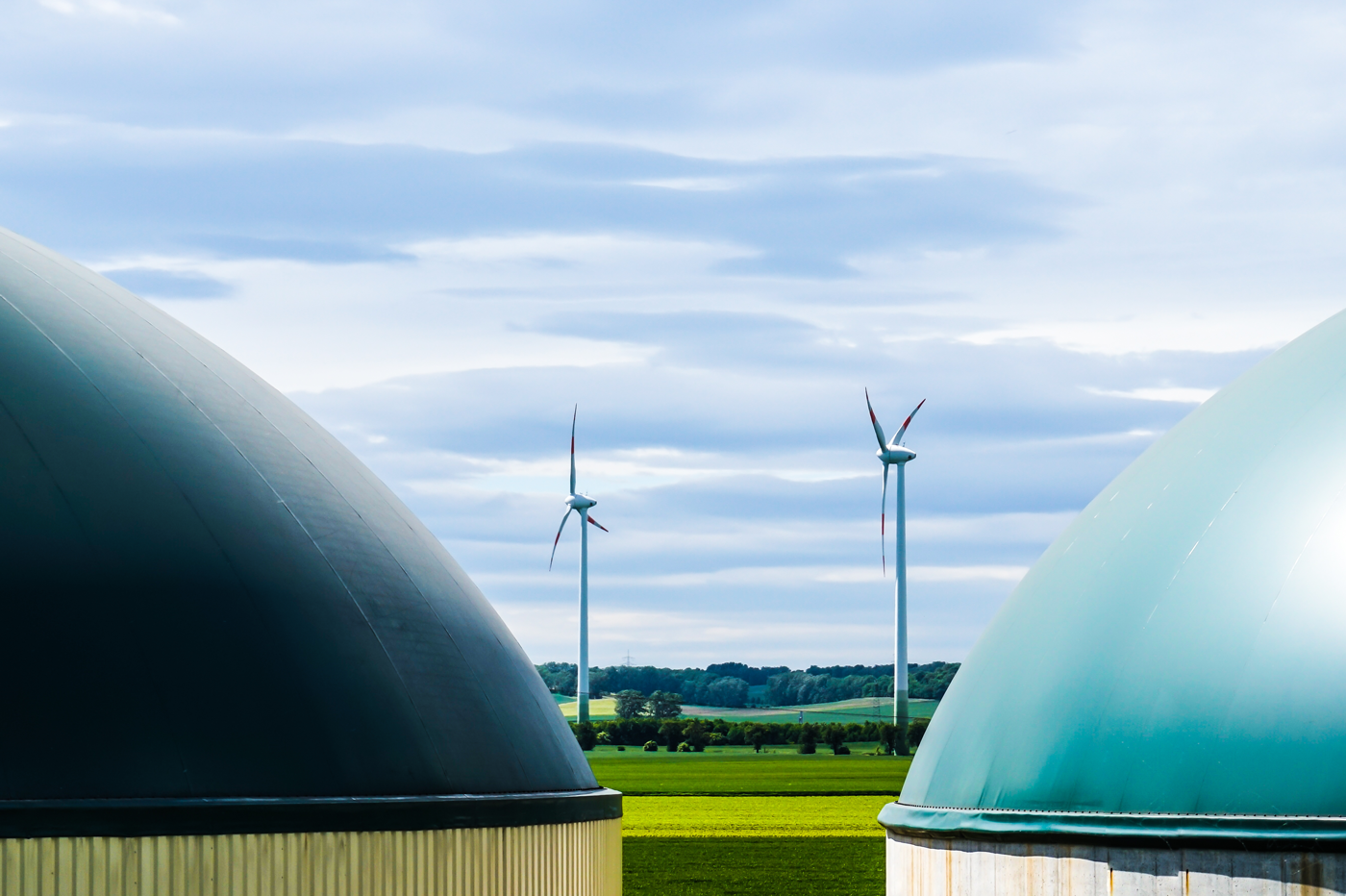 Two large dome-shaped structures of a biogas plant in the foreground, with three wind turbines in a green field and trees and hills in the background under a partly cloudy sky.