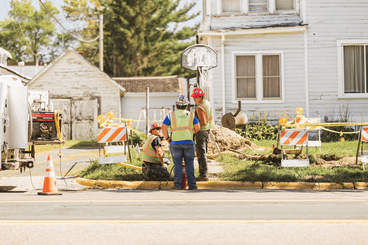 Construction workers in safety vests and helmets working on a site in front of a residential house, with construction equipment, traffic cones, caution tape, and barricades ensuring safety.