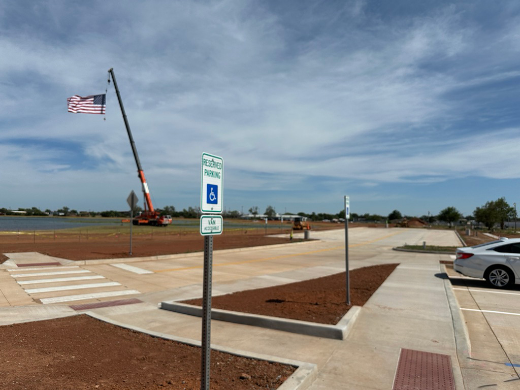 Construction site at Altus Reservoir with a crane lifting an American flag, adjacent to a parking lot with a reserved accessible parking sign and a white car parked nearby.