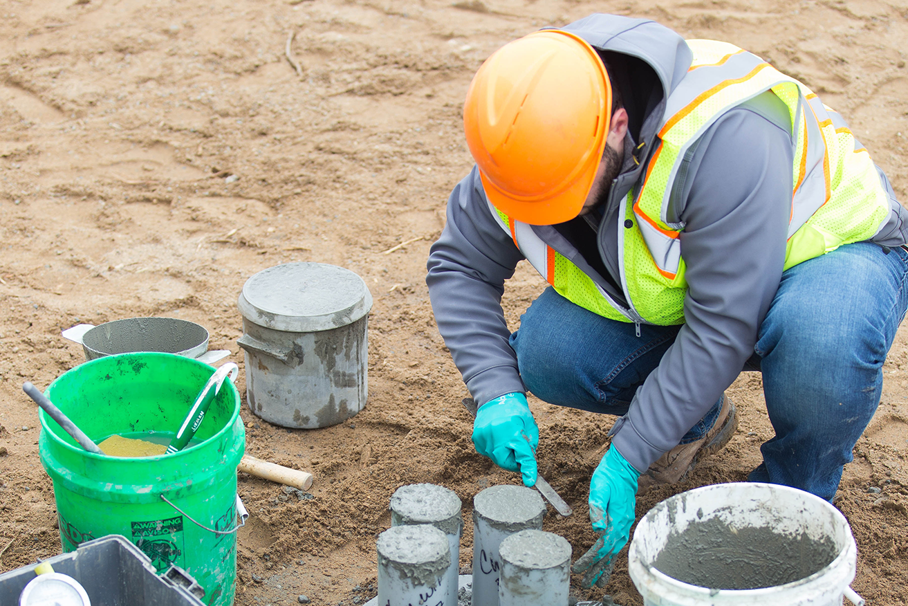 A construction worker wearing an orange hard hat, safety vest, and gloves kneels on sandy ground while preparing concrete test cylinders. Several buckets, tools, and partially filled concrete molds are arranged around the worker as they smooth the surface of the fresh concrete.