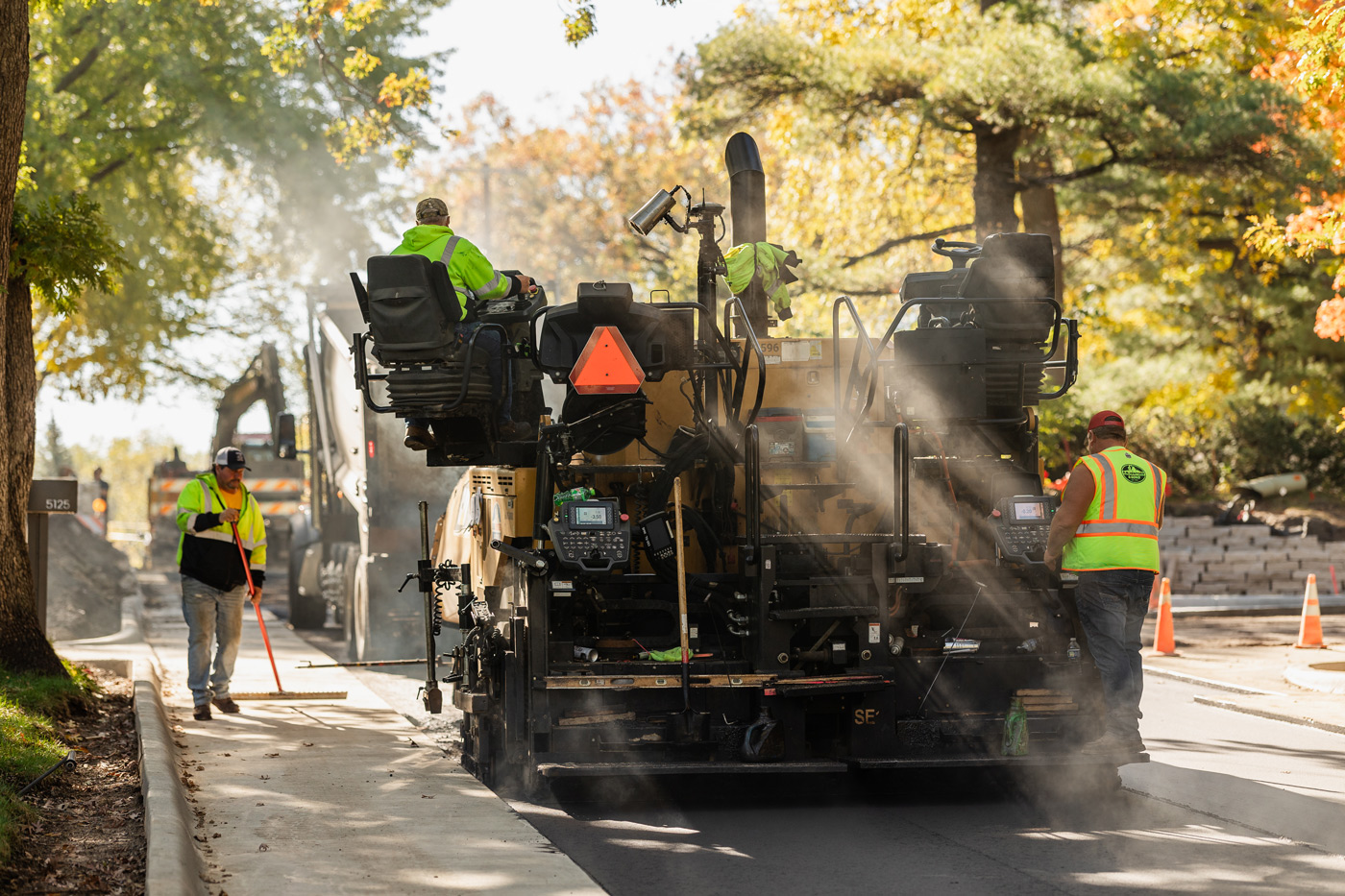 Road construction crew operates paving equipment and performs sidewalk work on an active street project.