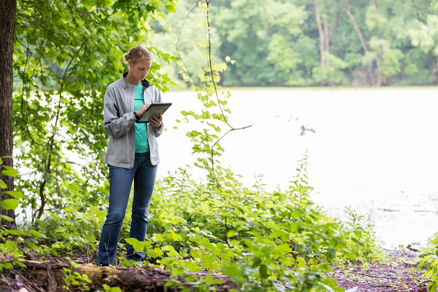 A field technician stands near dense vegetation by a lakeshore, using a tablet to document site conditions as part of utility damage prevention work.