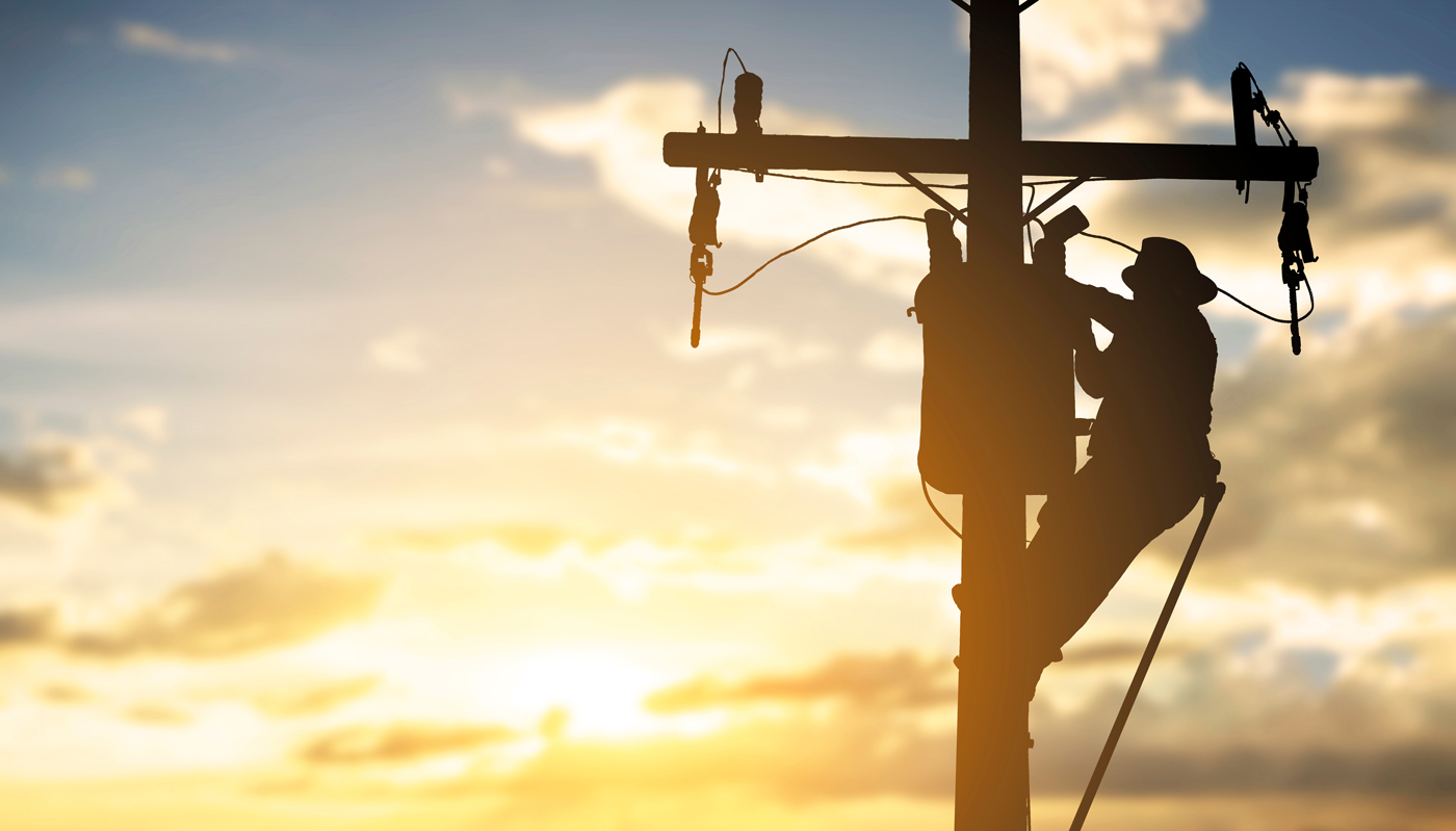 Silhouette of a utility worker climbing a distribution pole at sunset, working on pole‑top equipment and conductors. The scene highlights maintenance activities on overhead distribution infrastructure in low‑light conditions.