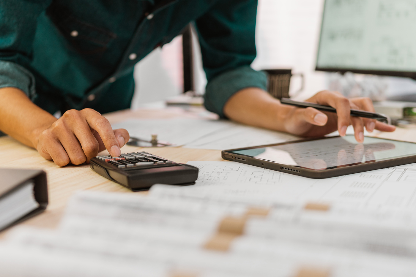 Close-up of a person performing calculation and estimation work at a desk, using a calculator and stylus while referencing engineering or architectural drawings. A tablet, printed plans, and project documents are spread across the workspace.