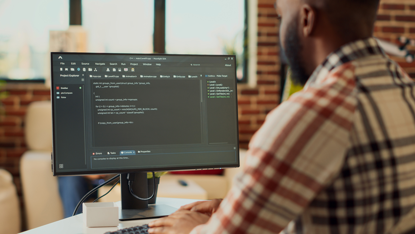 Person working at a desk with a computer monitor displaying software code in a modern office setting. The individual types on a keyboard while viewing a programming interface, with exposed brick walls and natural light in the background.