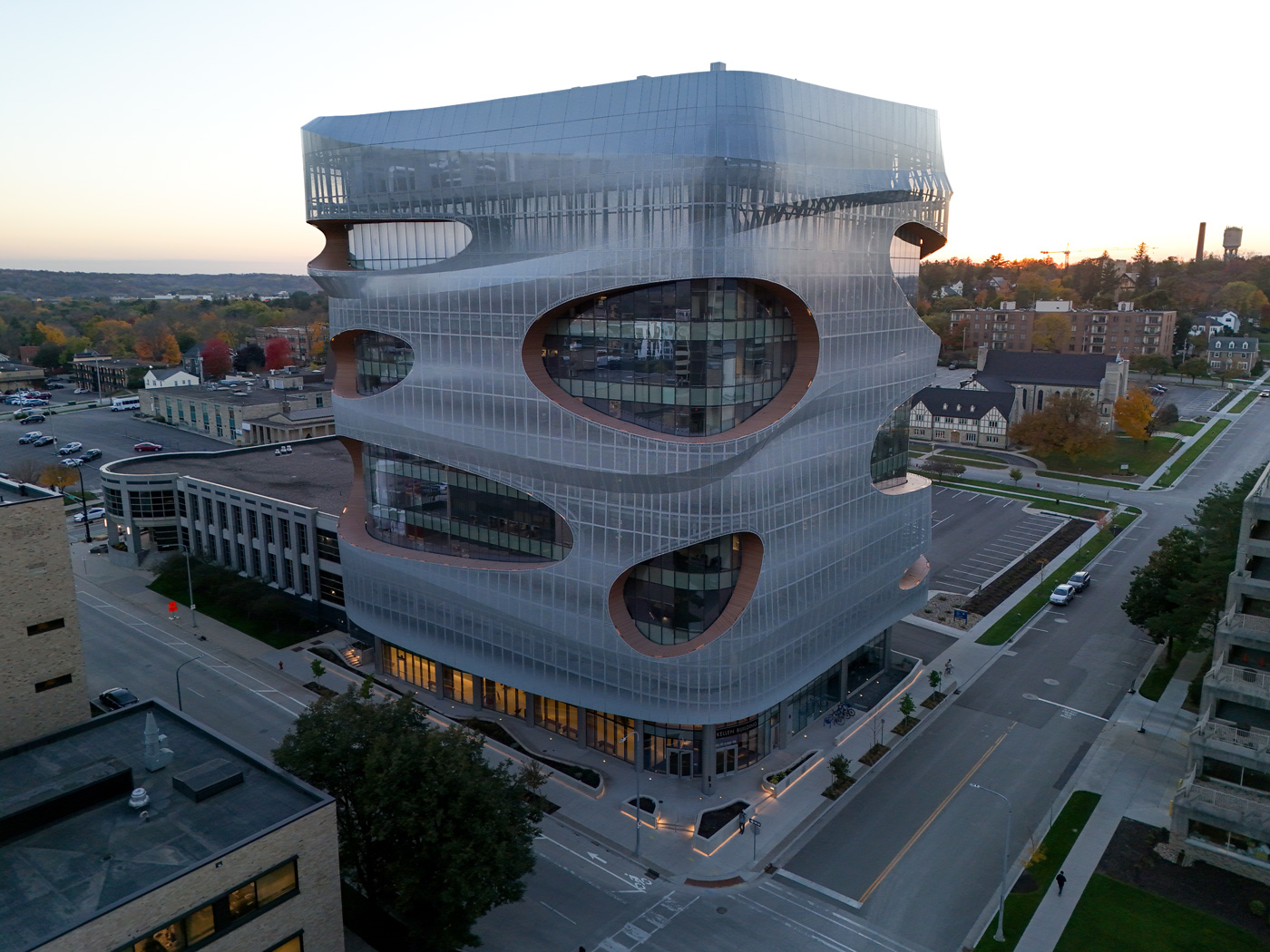 Futuristic building with curvy, perforated design, glass facade, and rounded edges at dusk. Surrounded by streets, trees, and a sunset-lit sky.