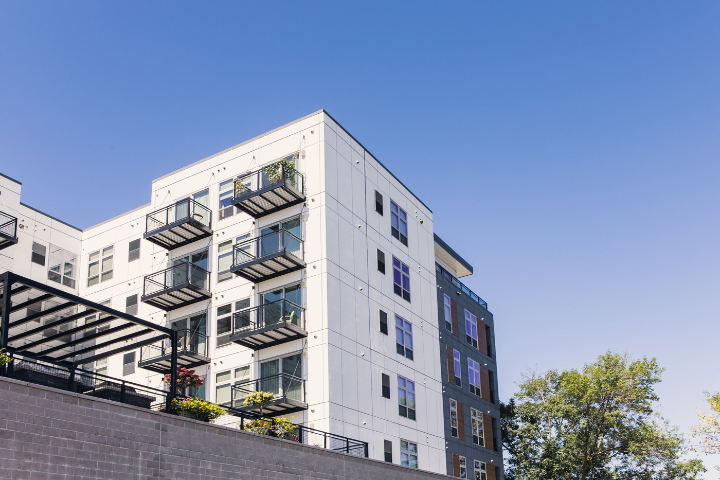 Modern apartment building with white and gray walls under a clear blue sky. Balconies with plants. Green trees in the background convey a serene atmosphere.