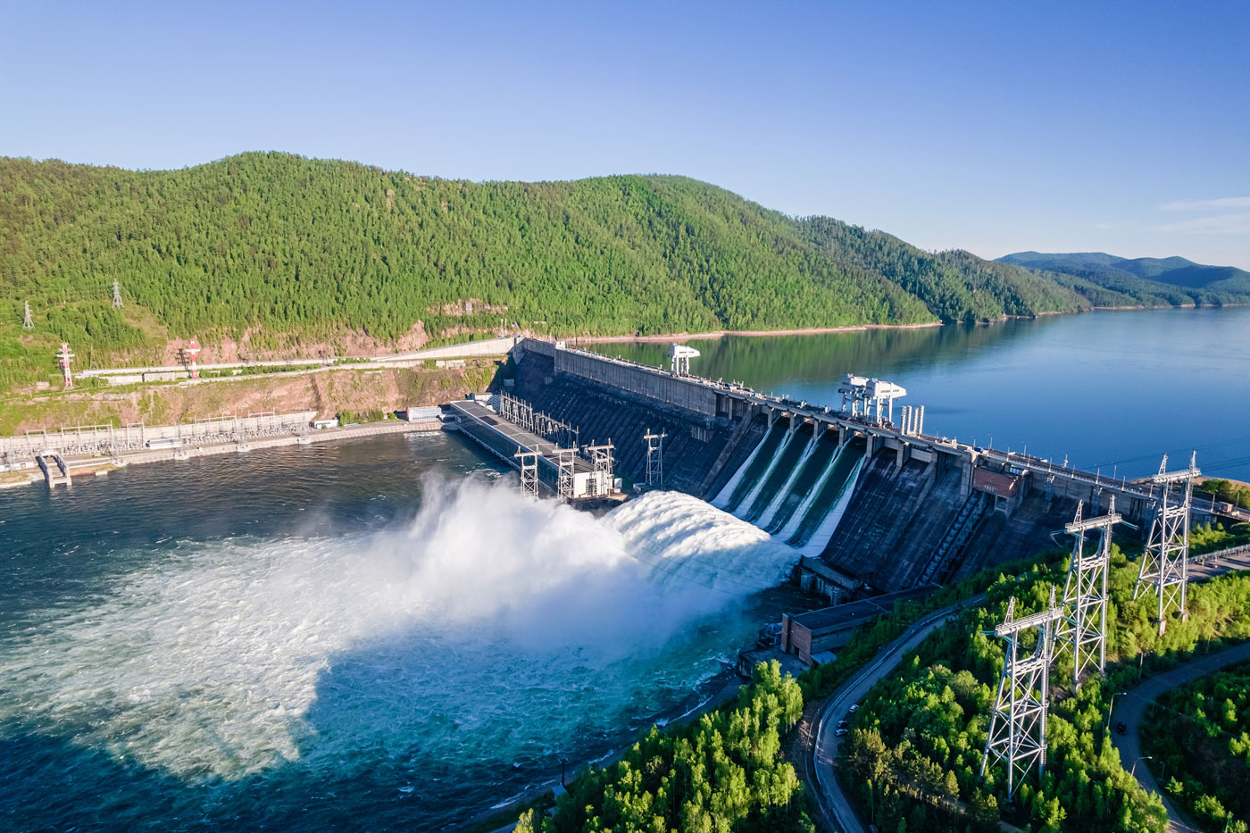 A large hydroelectric dam releasing water into a river, with lush green hills and a vast, calm reservoir under a clear blue sky. The scene is vibrant and dynamic.