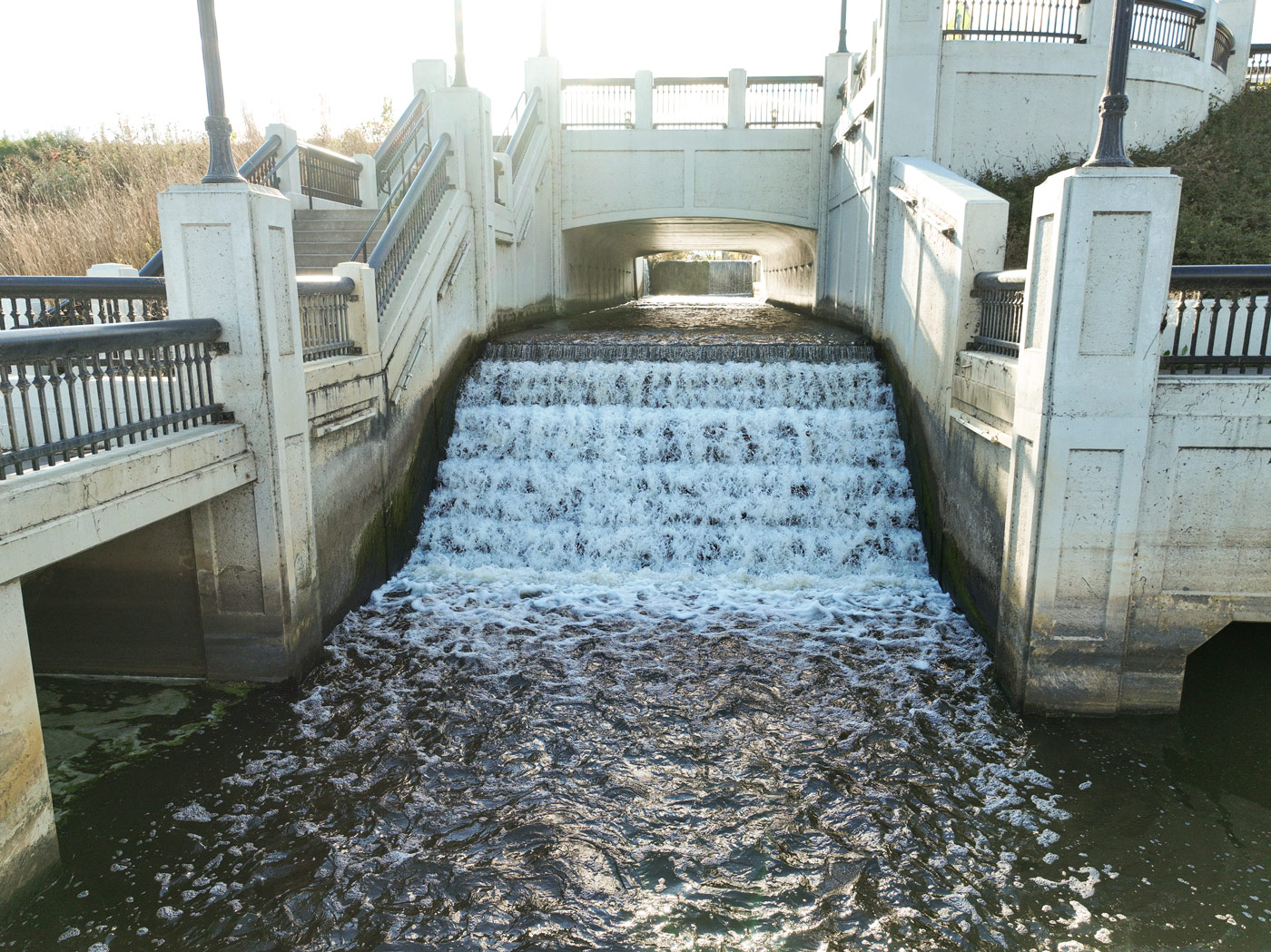 Water flows down a stepped concrete spillway beneath a pedestrian bridge.