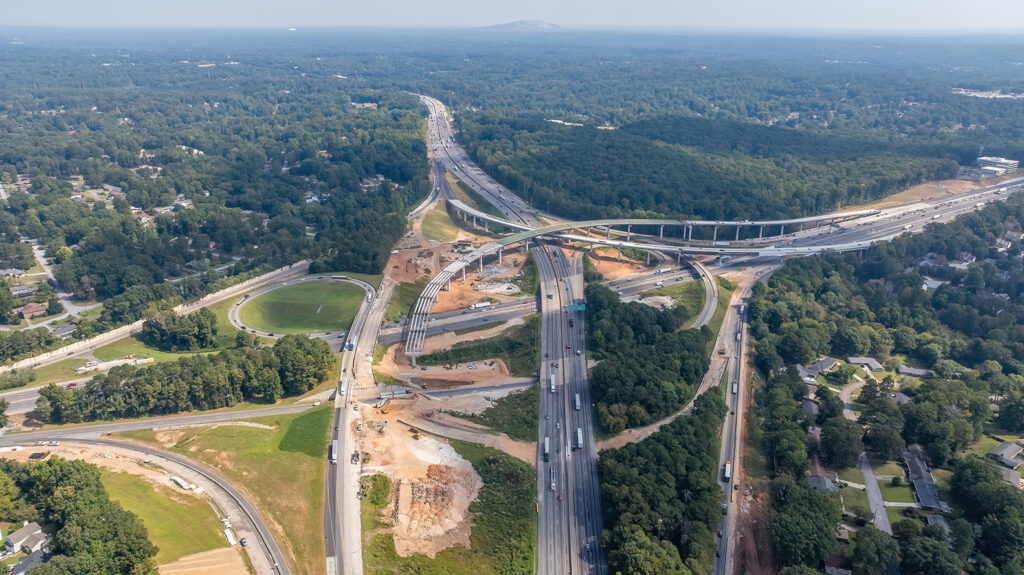 A high-angle aerial view of a large highway interchange under construction, surrounded by dense green forest and suburban neighborhoods. Multiple elevated ramps and bridges cross over one another, with sections of roadway being widened or rebuilt. Construction equipment and cleared earth are visible along the interchange. Traffic moves along the completed portions of the highway, and a distant tree‑covered hill rises on the horizon under a hazy sky.