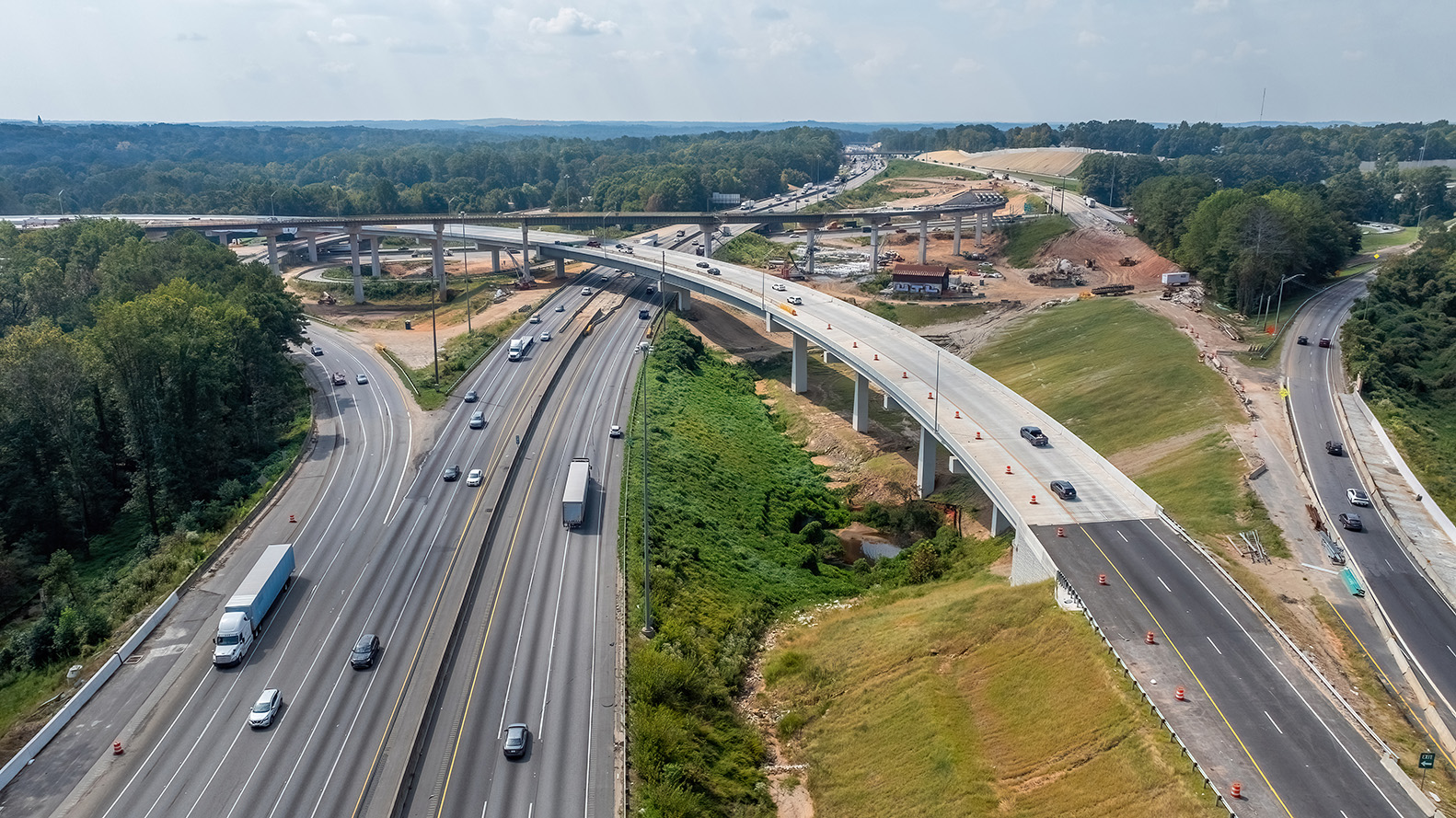 A mid-level aerial view of an active highway interchange construction site. Multiple elevated ramps curve over a busy multi‑lane interstate, with traffic moving in both directions. Several bridges are newly built or partially completed, surrounded by earthwork, construction equipment, and cleared areas. Dense green trees frame the scene, with rolling hills visible in the distance under a hazy sky.