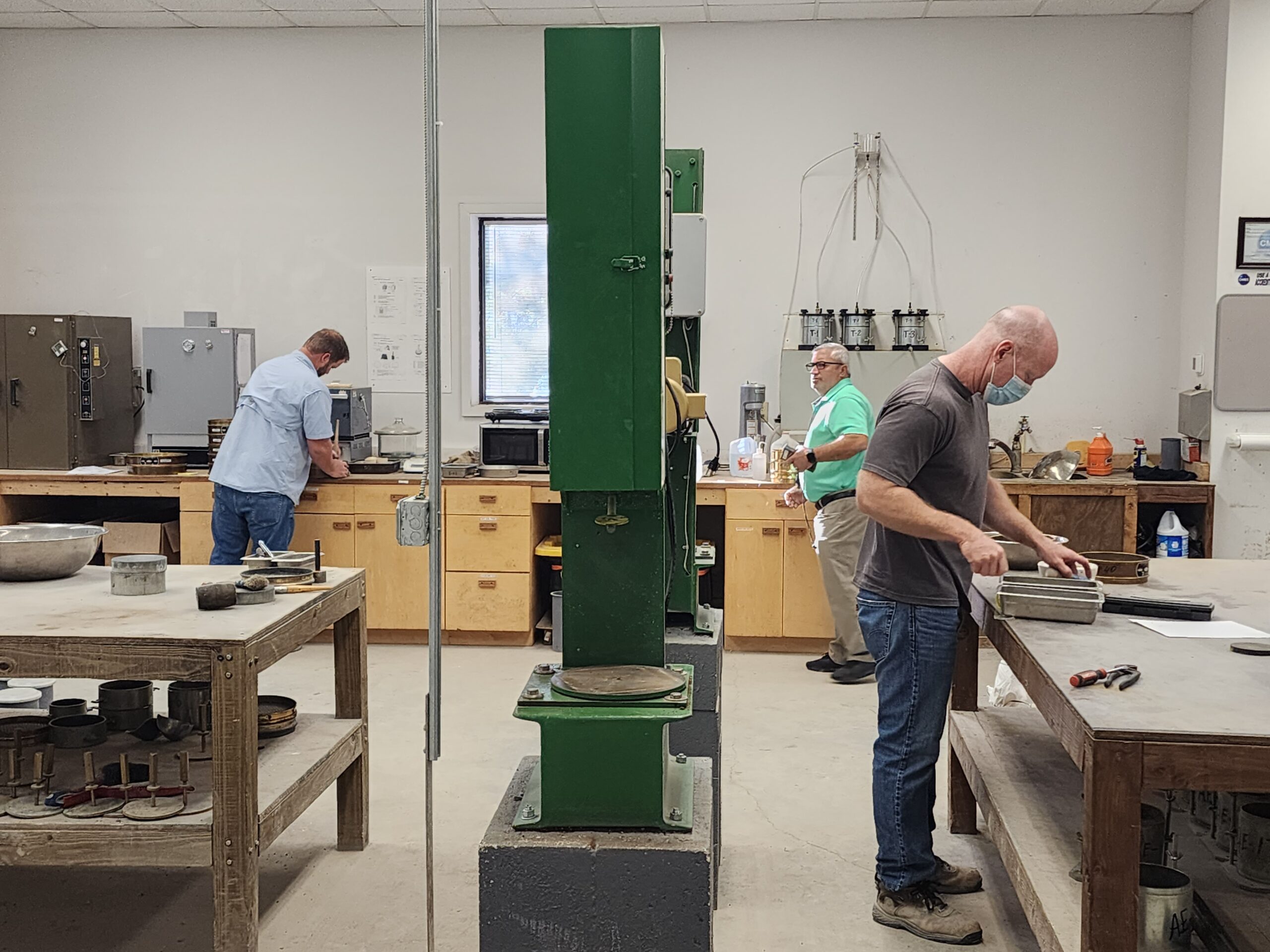 A laboratory workspace with three people engaged in material testing tasks at separate workstations. The room contains large industrial equipment, metal sample containers, and wooden workbenches. A tall green testing machine stands in the center of the space, dividing the room. Shelving units, ovens, and tools line the back wall, contributing to an organized, functional lab environment.
