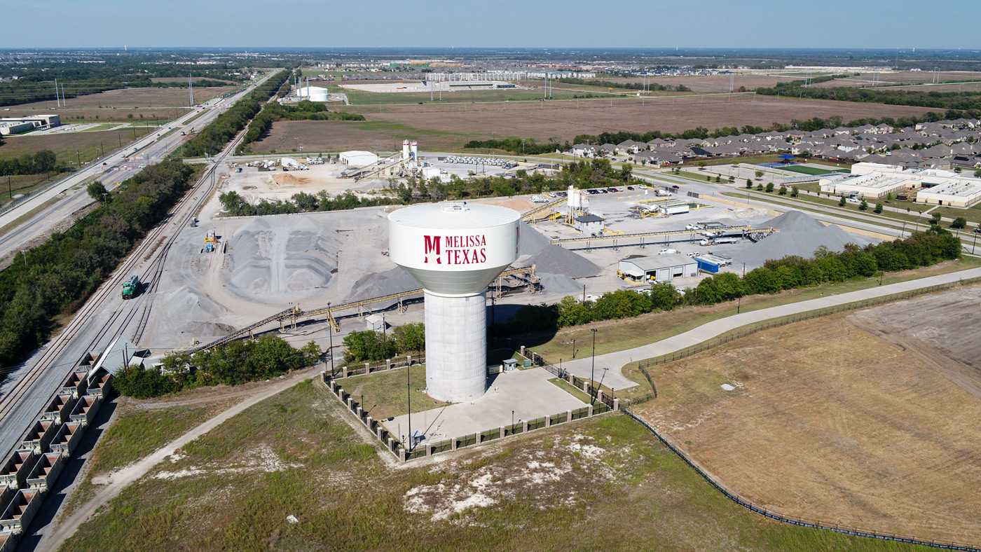 Aerial view of a water tower labeled "Melissa, Texas" near an industrial area with construction materials. Nearby are railway tracks and a highway.