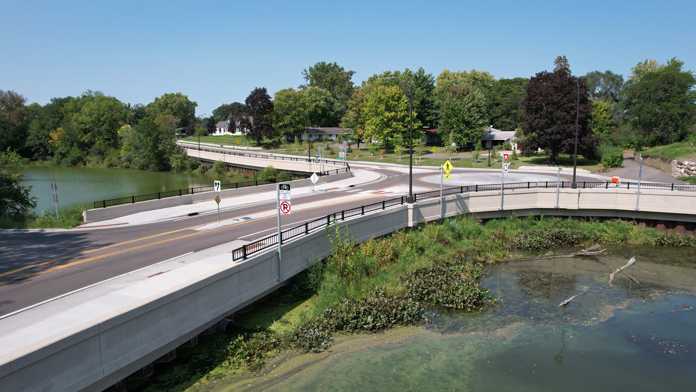 Curved bridge over a lush green area beside a small body of water, with residential houses and trees in the background under a clear blue sky.