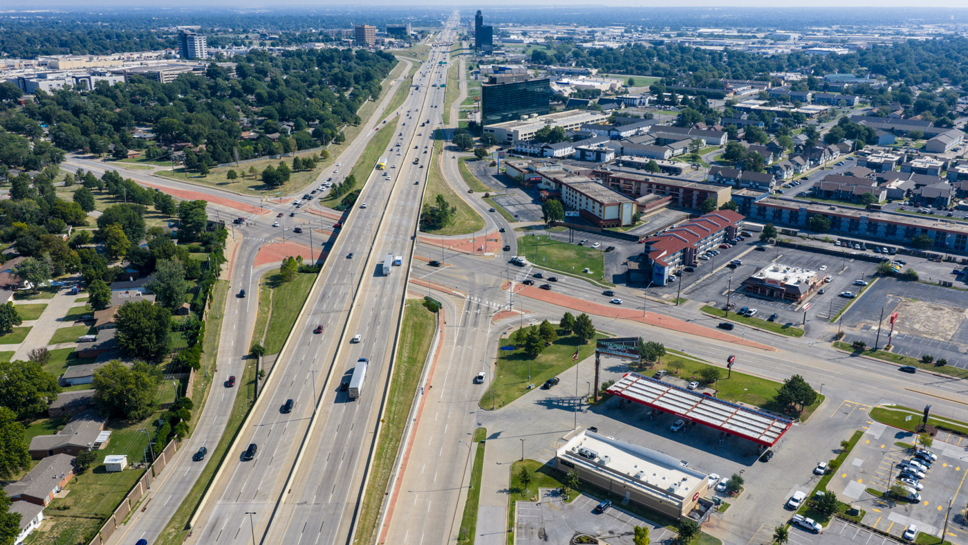 Aerial view of a busy multi-lane highway intersection in an urban area, surrounded by commercial buildings and green spaces. Vehicles are visible on the road.