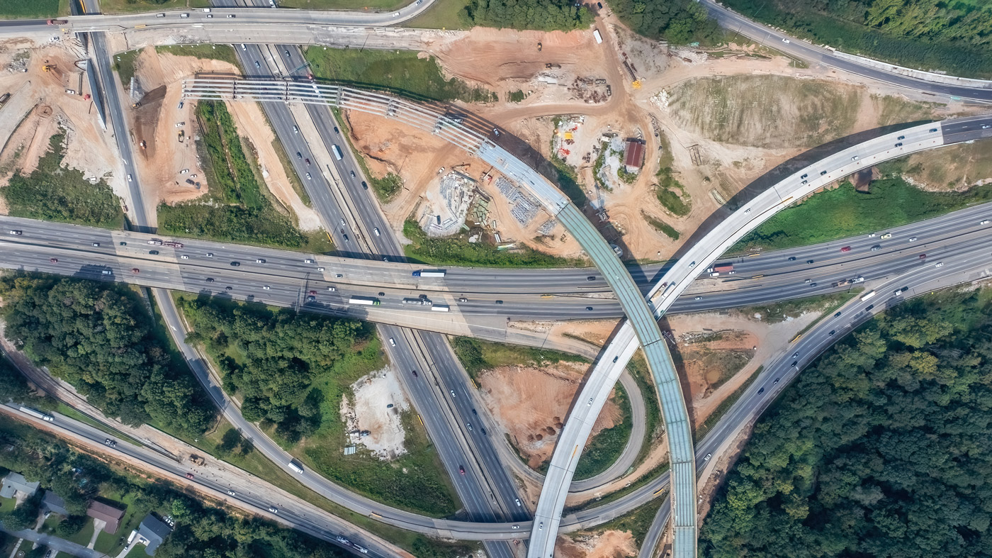 Aerial view of a complex highway interchange surrounded by greenery and construction sites. Multiple overpasses and roads intersect, conveying a sense of urban development and movement.