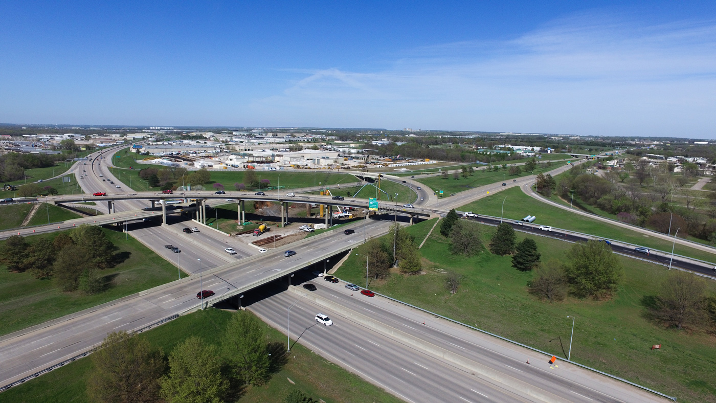Aerial view of a highway interchange with multiple overpasses and cars. Surrounding landscape includes green spaces, trees, and industrial buildings under a clear blue sky.