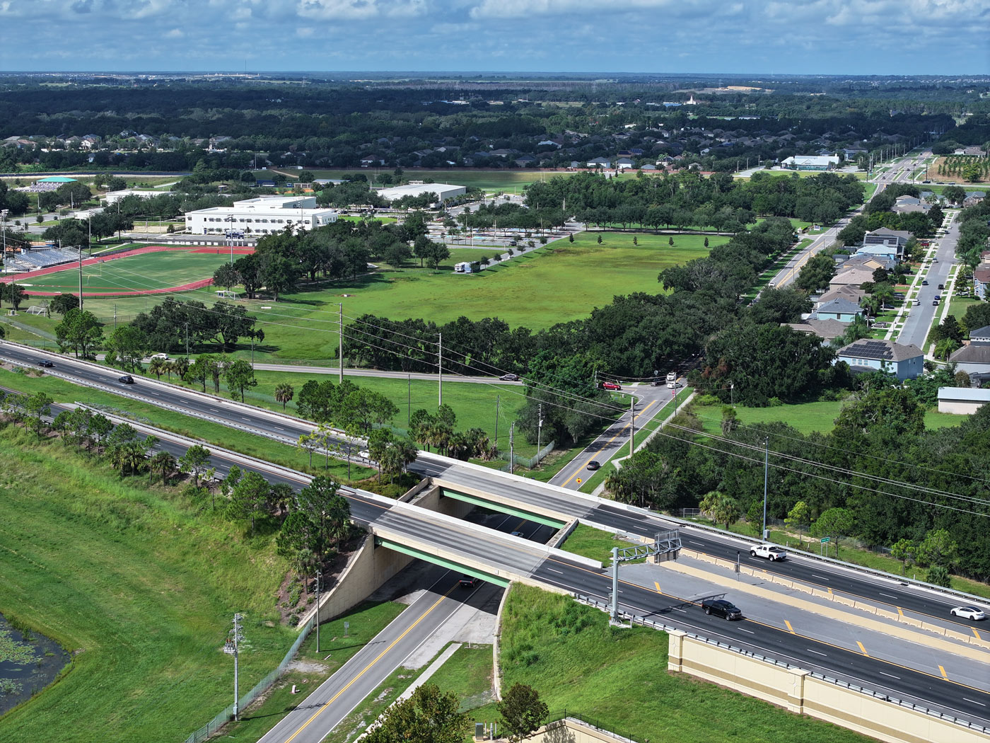 Aerial view of a busy highway intersection in a suburban area with green fields, trees, and scattered buildings. Blue sky and scattered clouds above.
