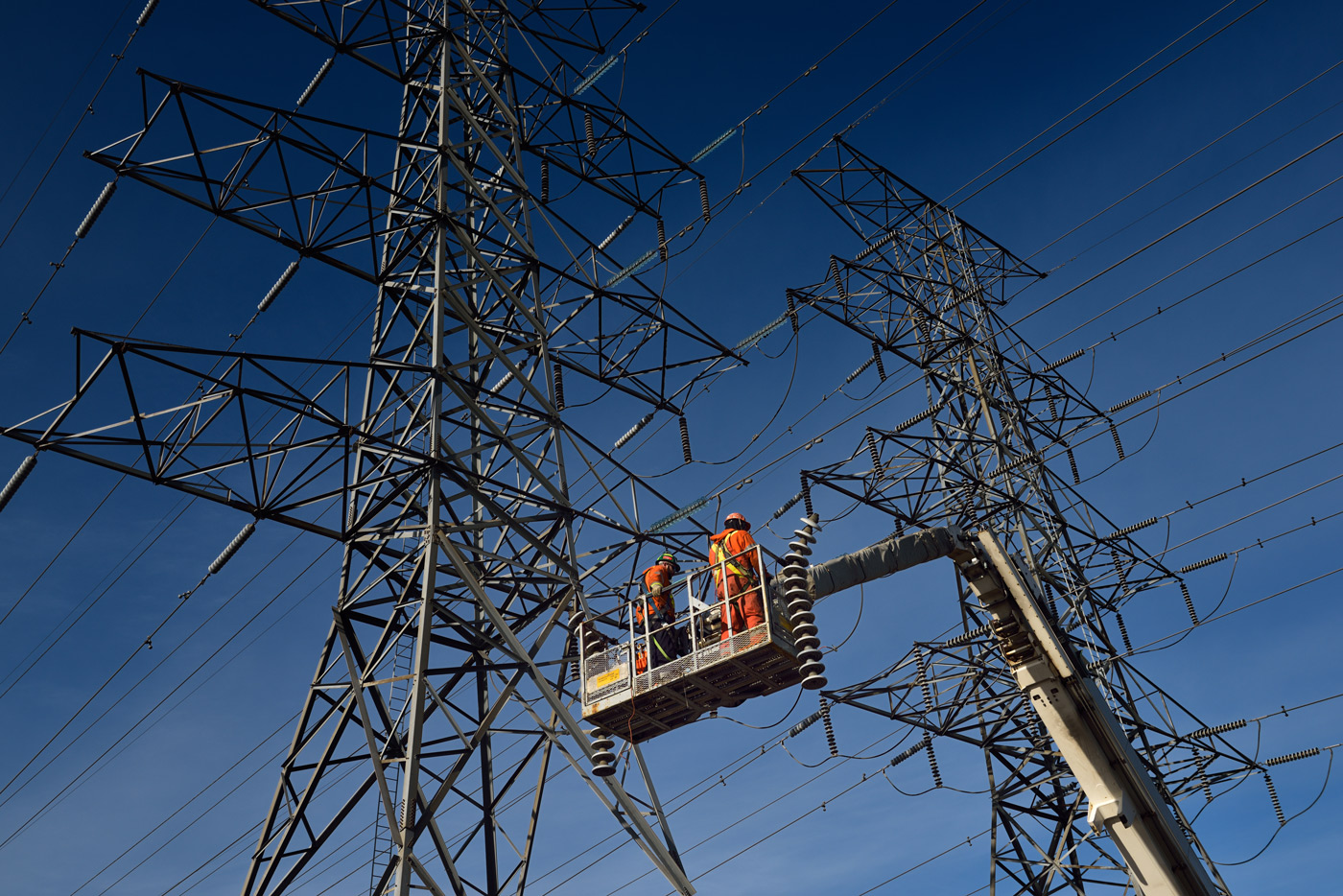 Utility workers in a lift perform maintenance on high‑voltage transmission lines beneath steel lattice towers.