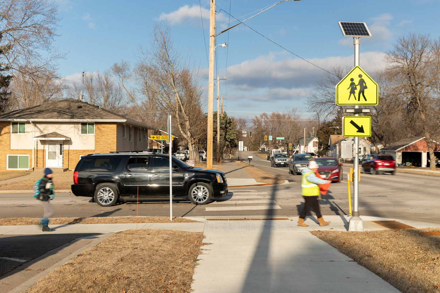 A sunny street scene shows a child with a backpack crossing a road, assisted by a crossing guard holding a flag. Nearby, a bright yellow pedestrian sign is visible, while cars wait on the street.
