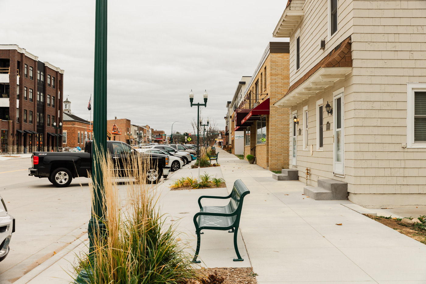 Small-town street with parked cars, a lone green bench, and buildings on a cloudy day. The scene conveys quiet, everyday life in the town.