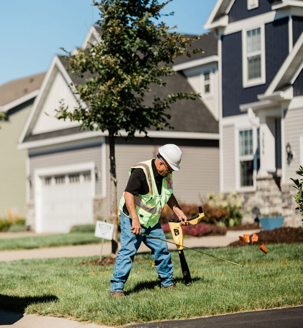 A construction worker in a safety vest and hard hat measures a front lawn in a suburban neighborhood, suggesting utility work on a sunny day.