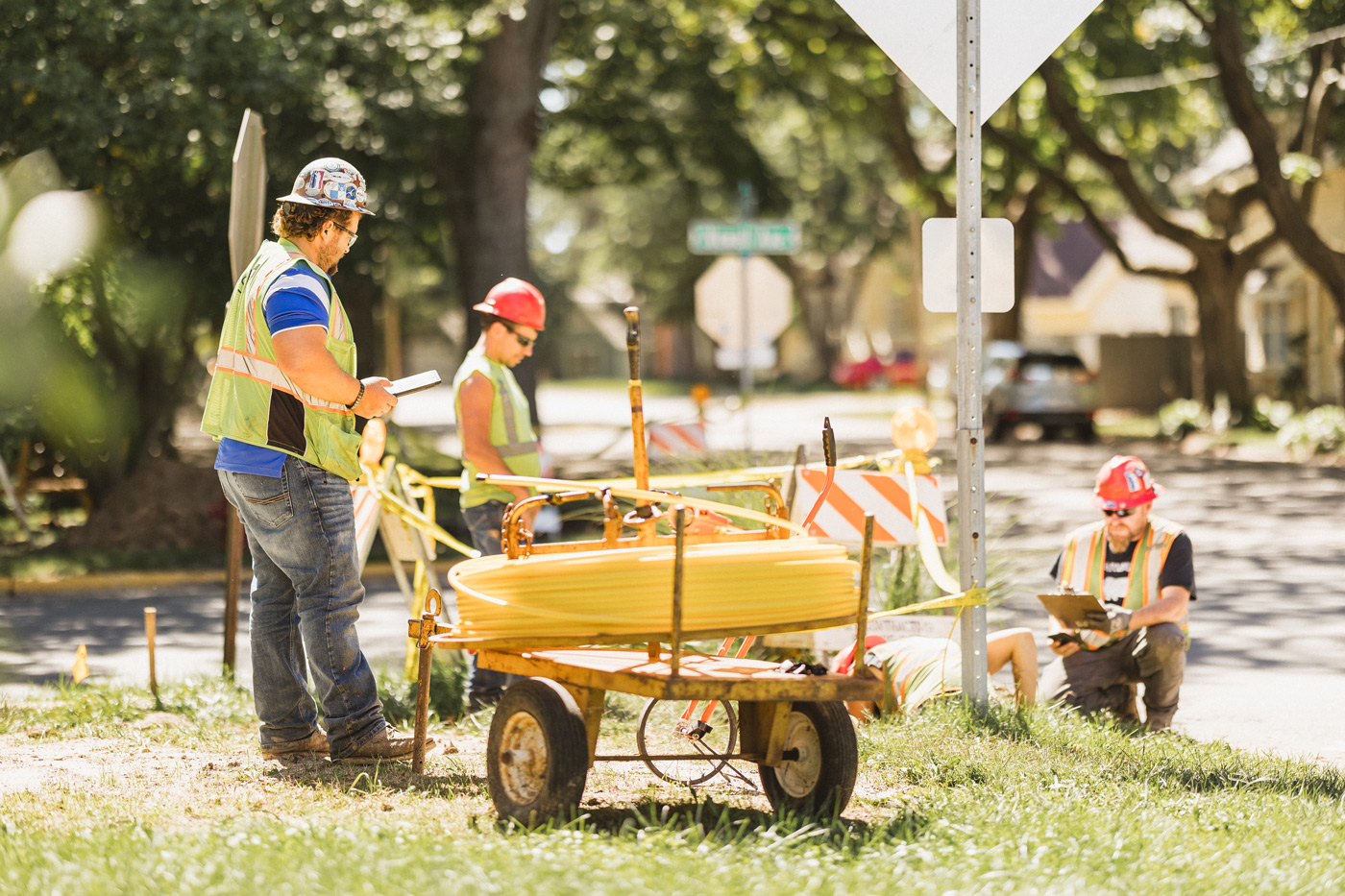 Construction workers in safety vests and hard hats work on a sunny street corner. One reads a tablet, surrounded by equipment and orange barriers.