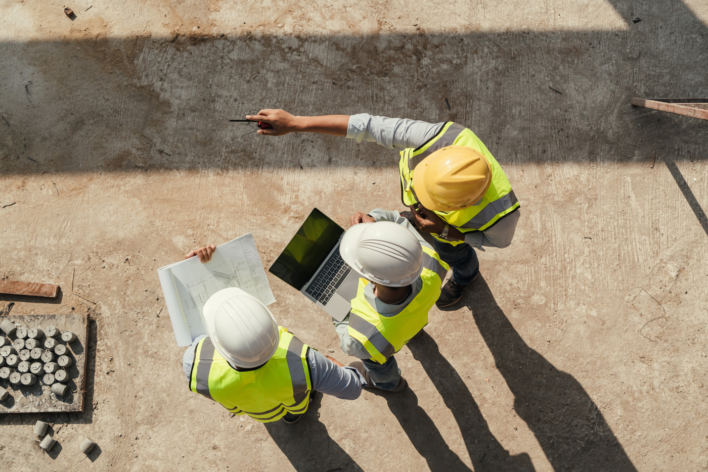 Aerial view of three construction team members wearing hard hats and high‑visibility vests reviewing structural plans and data on a laptop at an active jobsite. One team member points toward site features while others study design drawings, illustrating collaborative field assessment and project coordination.