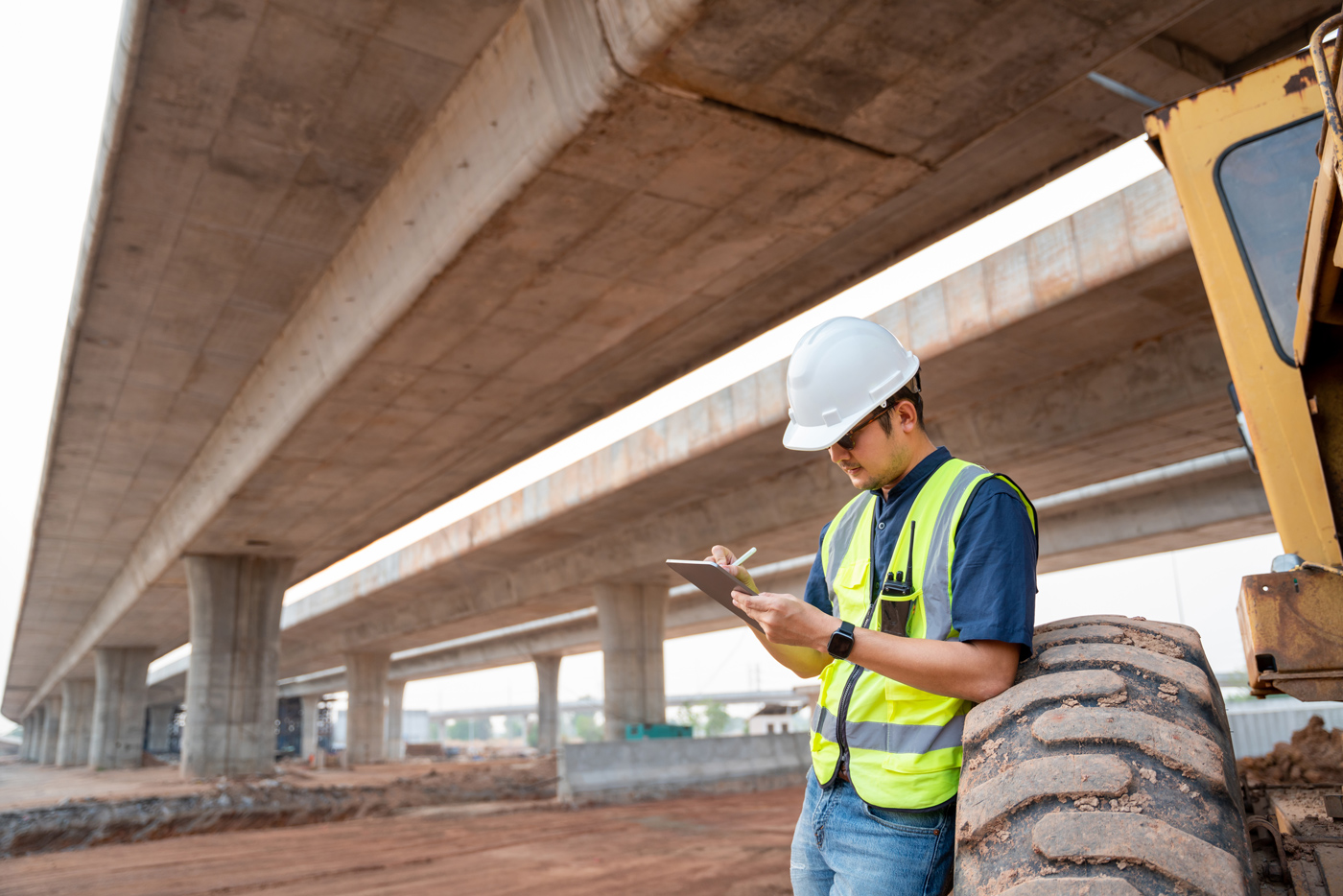 Construction inspector wearing a hard hat and high‑visibility vest documenting site conditions on a clipboard while standing beside heavy equipment under an elevated roadway structure. The scene shows active civil infrastructure work with bridge supports, grading, and earthwork in progress.