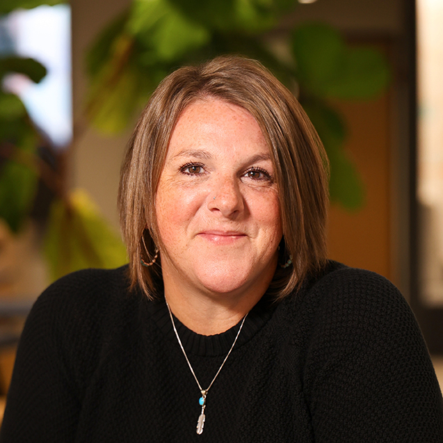 Portrait of Allison Whitsitt in a black sweater and a silver necklace with a turquoise pendant, seated indoors with green plants in the background.