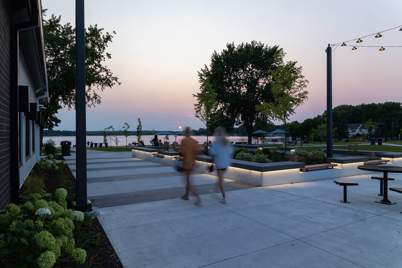People walking through a lakeside park plaza at sunset with illuminated seating, trees, and water in the background.