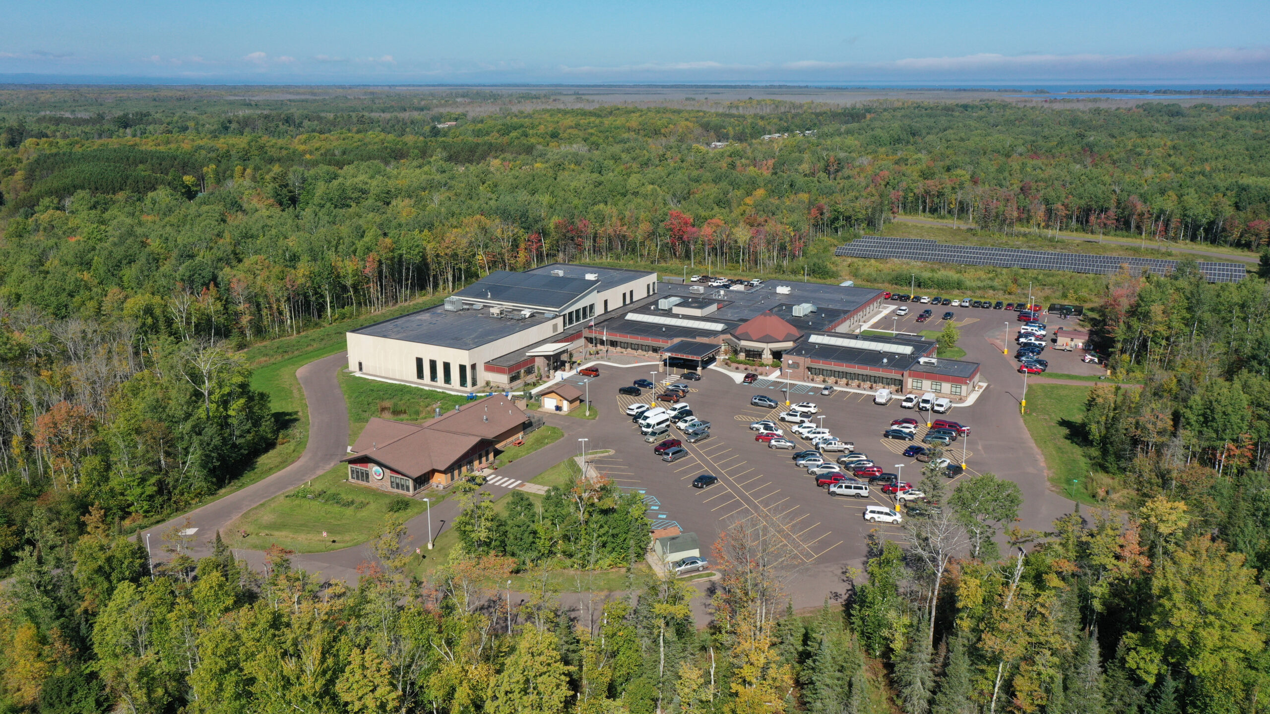 Aerial view of a campus with multiple buildings and parking areas surrounded by dense forest, with vehicles visible and open land extending to the horizon.
