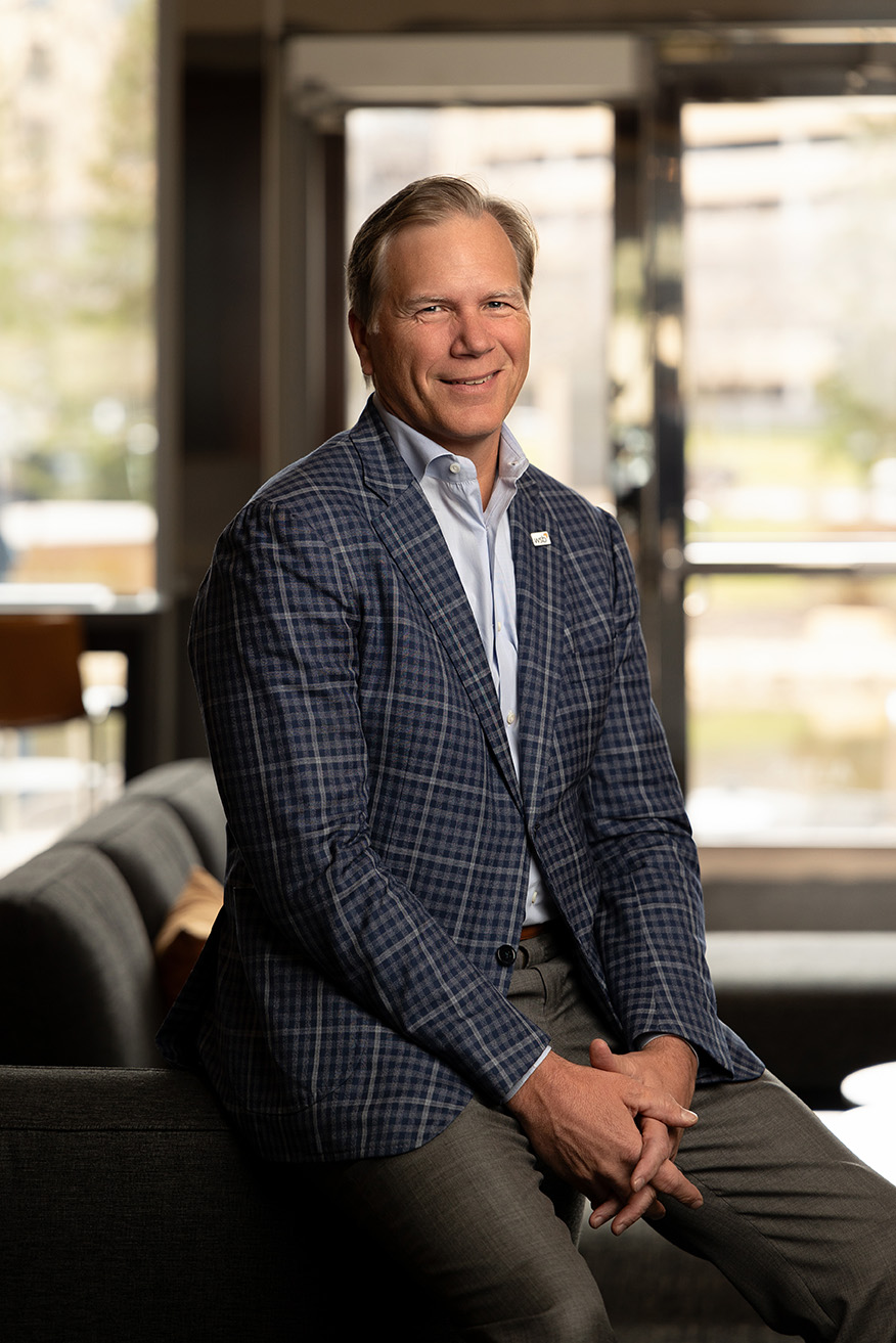 A person wearing a blue checkered blazer, white dress shirt, and gray pants is seated on the arm of a sofa in a modern indoor setting. Their hands are loosely clasped, and large windows behind them reveal an outdoor scene with soft, natural light.