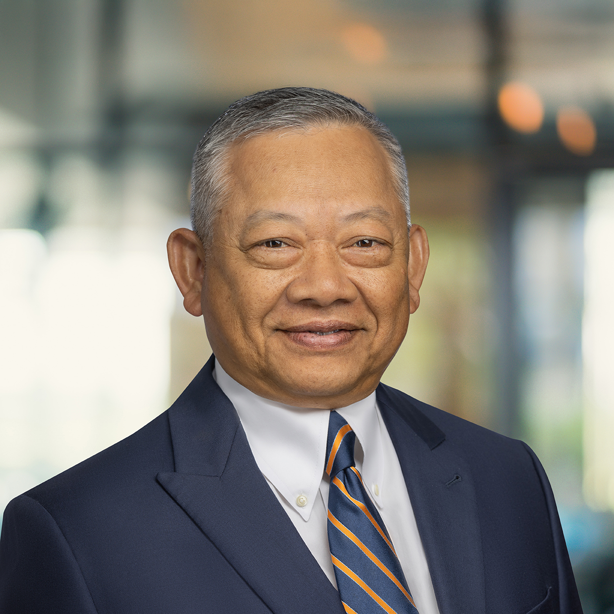 Professional head-and-shoulders portrait of Channy Soeur wearing a navy suit jacket, white dress shirt, and striped tie, photographed indoors in a modern office setting with large windows and soft, natural lighting.