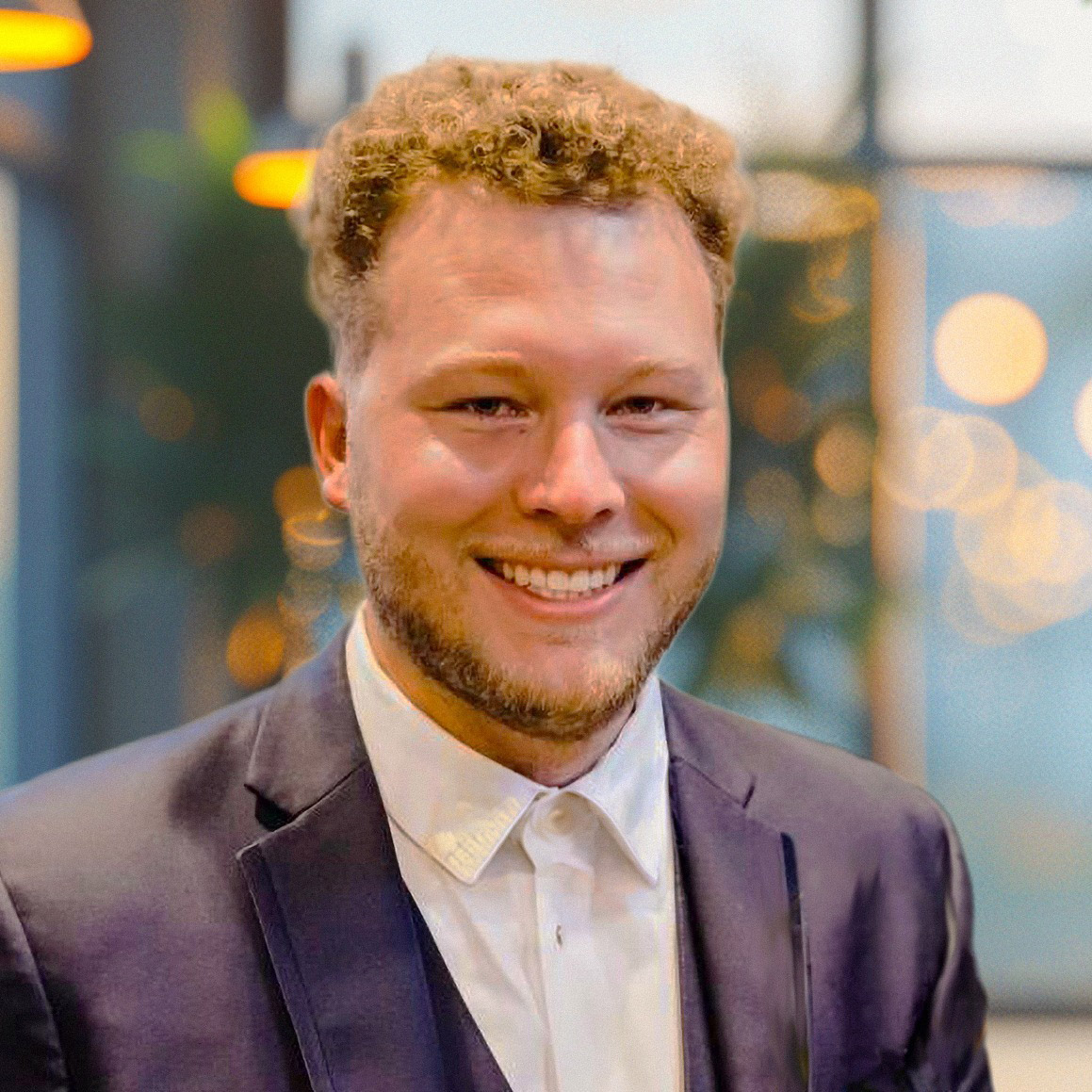 Cody Hans wearing a dark blazer and white button-down shirt, standing indoors with a softly lit background featuring warm lights and large windows.