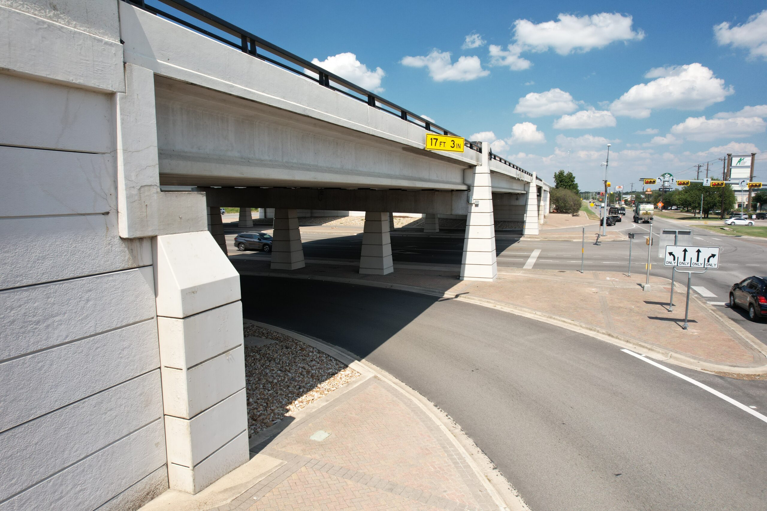 Ground‑level view beneath an elevated highway bridge showing support columns and a curved roadway passing underneath. Cars travel through a wide intersection in the background under a sunny sky.