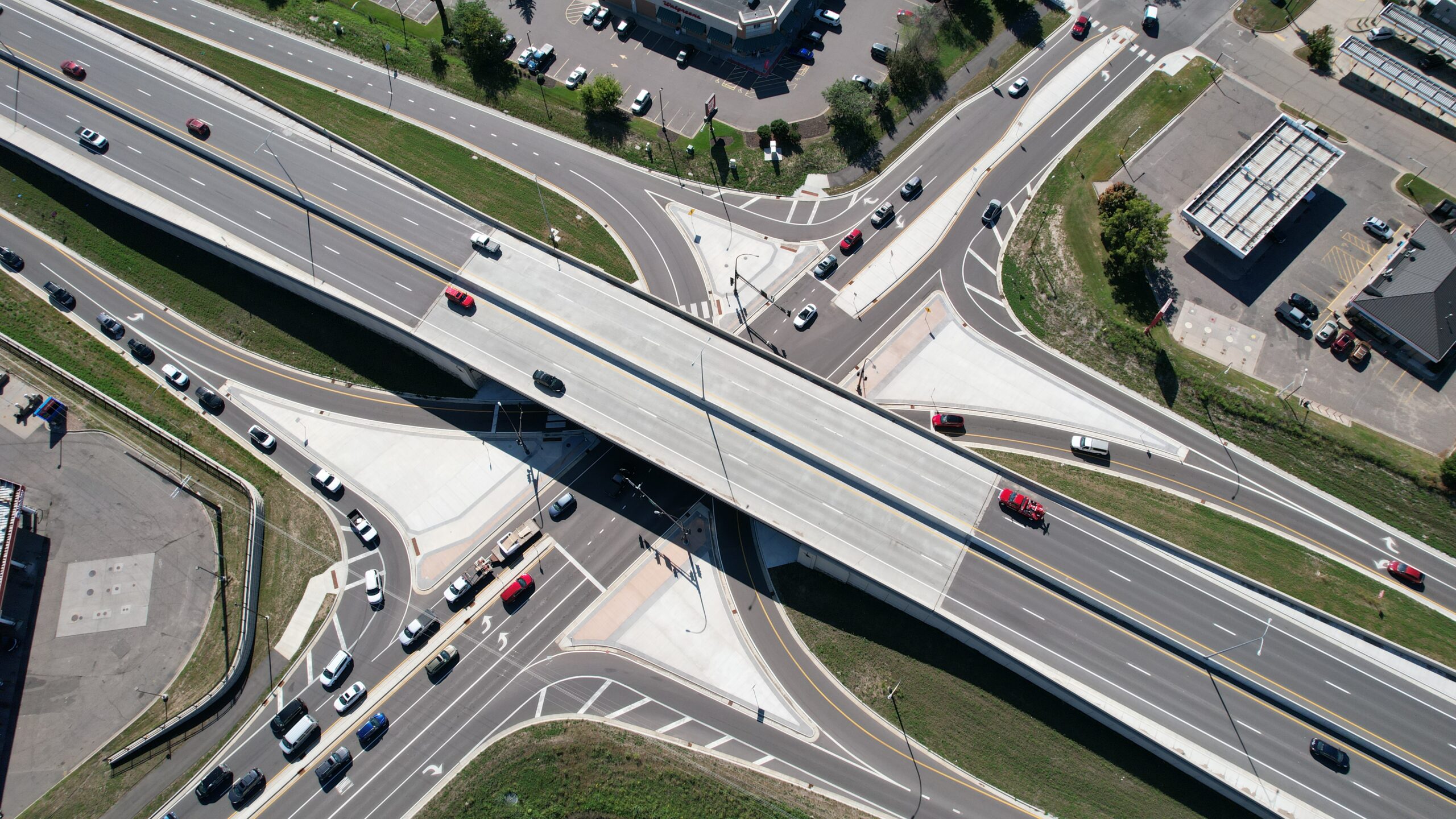 Aerial overhead view of a highway interchange with an elevated overpass crossing above multiple surface streets. Cars travel in both directions on the overpass and on the connecting roads below. The interchange includes curved ramps, turn lanes, and traffic signals. Surrounding the roads are commercial buildings, parking lots, and green spaces.