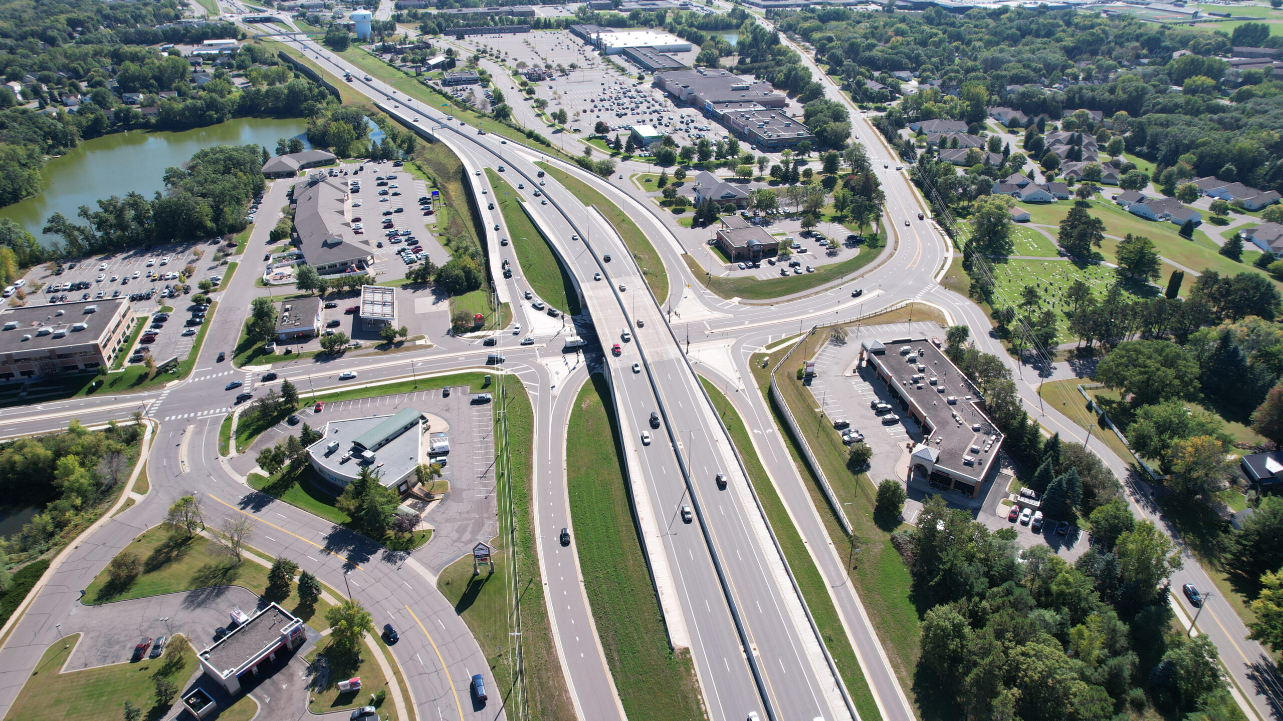 Aerial view of a large highway interchange featuring an overpass with multiple lanes of traffic. Several curved ramps connect surrounding roads to the main highway. Commercial buildings, parking lots, and office complexes border the interchange, with residential neighborhoods and green spaces visible in the distance. A small lake and clusters of trees appear on the left side of the image.