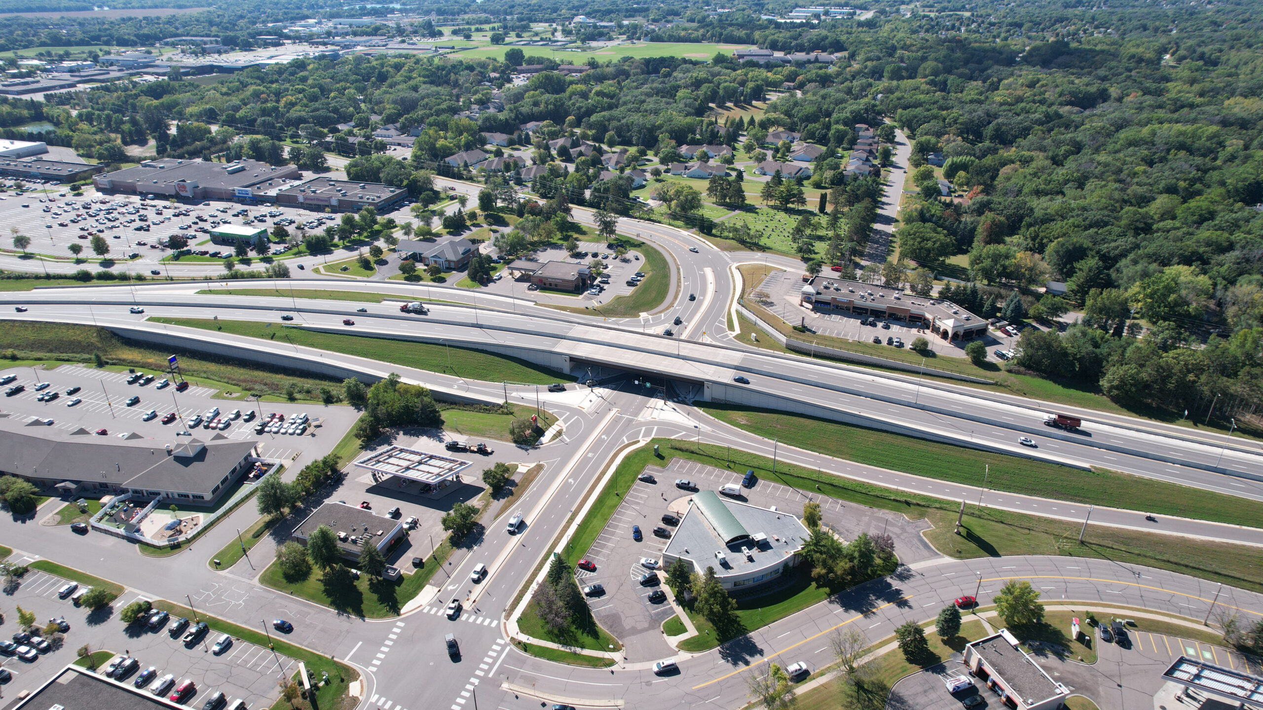 Aerial view of a highway interchange with an overpass running left to right across the image. Multiple lanes of traffic flow in both directions, with curved ramps connecting to surface streets below. Surrounding the interchange are commercial buildings, parking lots, and tree-lined neighborhoods. A large wooded area fills the right side of the image, while open fields and developed areas extend into the distance.