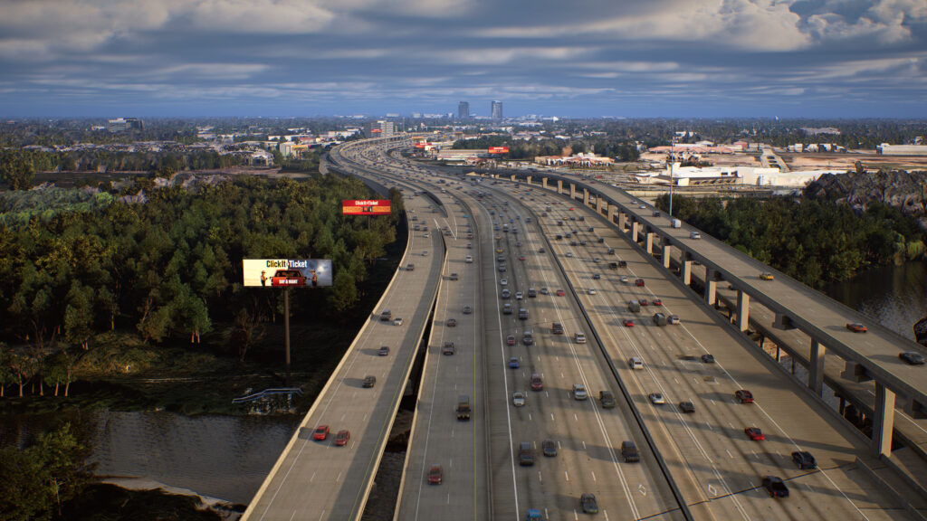 Wide freeway filled with heavy traffic in both directions. Elevated lanes run parallel on the right, with billboards and wooded areas below and a distant city skyline under a cloudy sky.