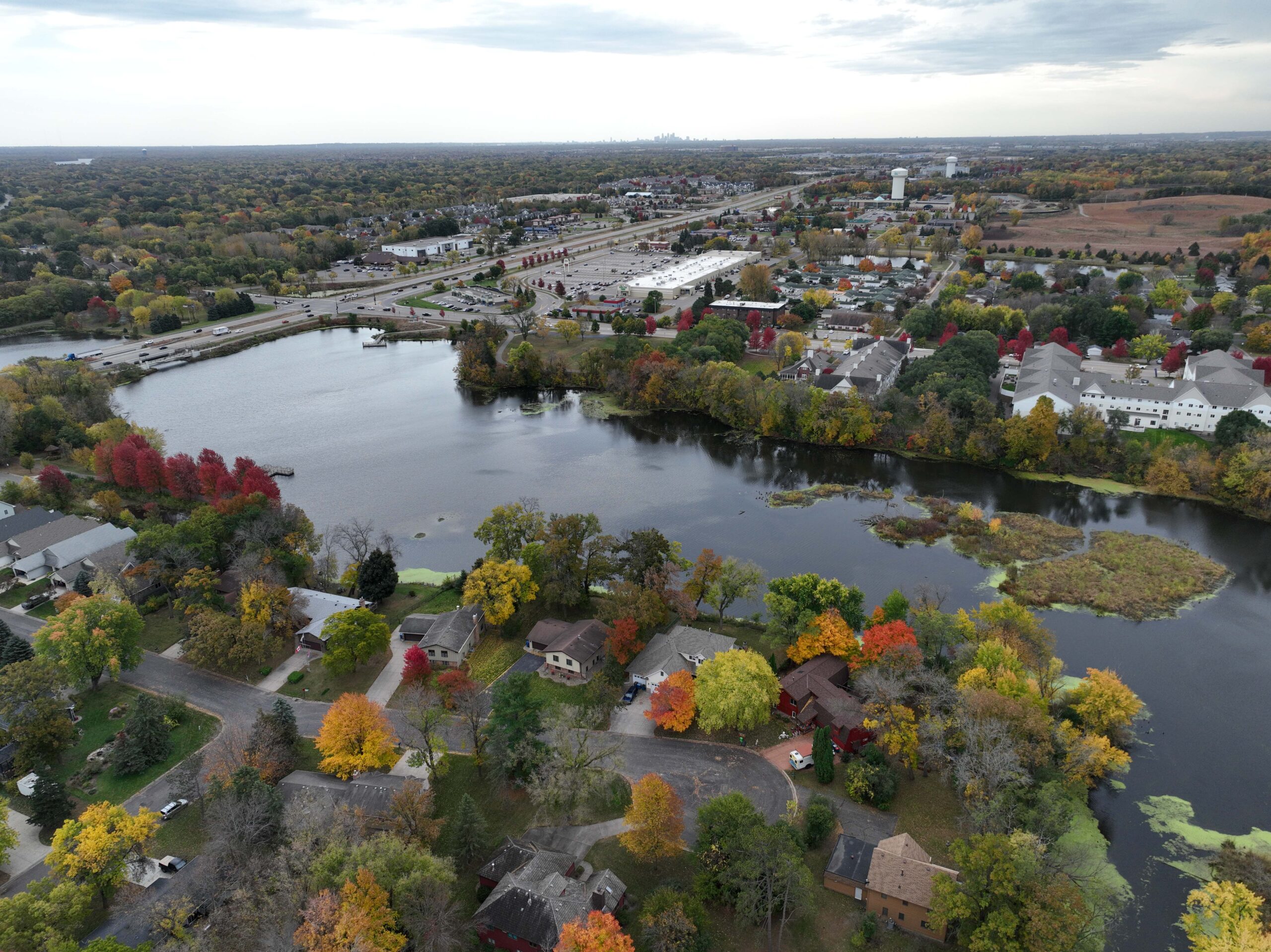 Aerial view of a lake bordered by residential homes, tree‑lined streets, and a nearby commercial corridor in autumn.