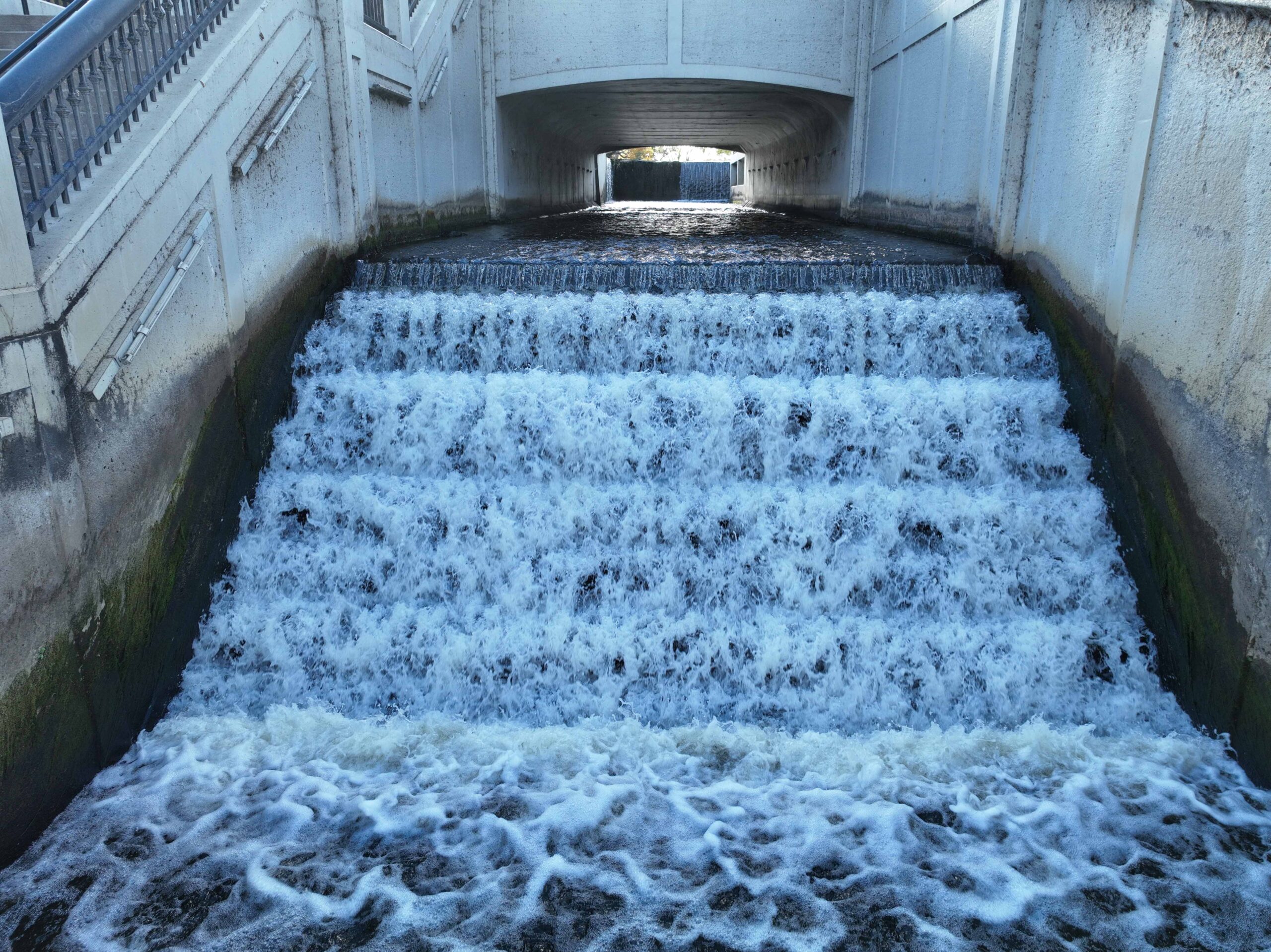 Water flowing over a stepped concrete spillway beneath a roadway structure.