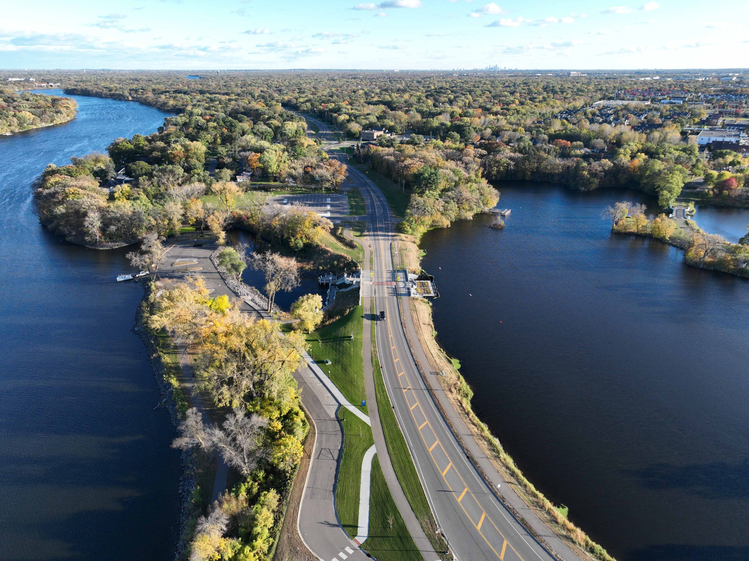 Aerial view of a roadway corridor between two lakes, with surrounding trees, trails, and open green space.
