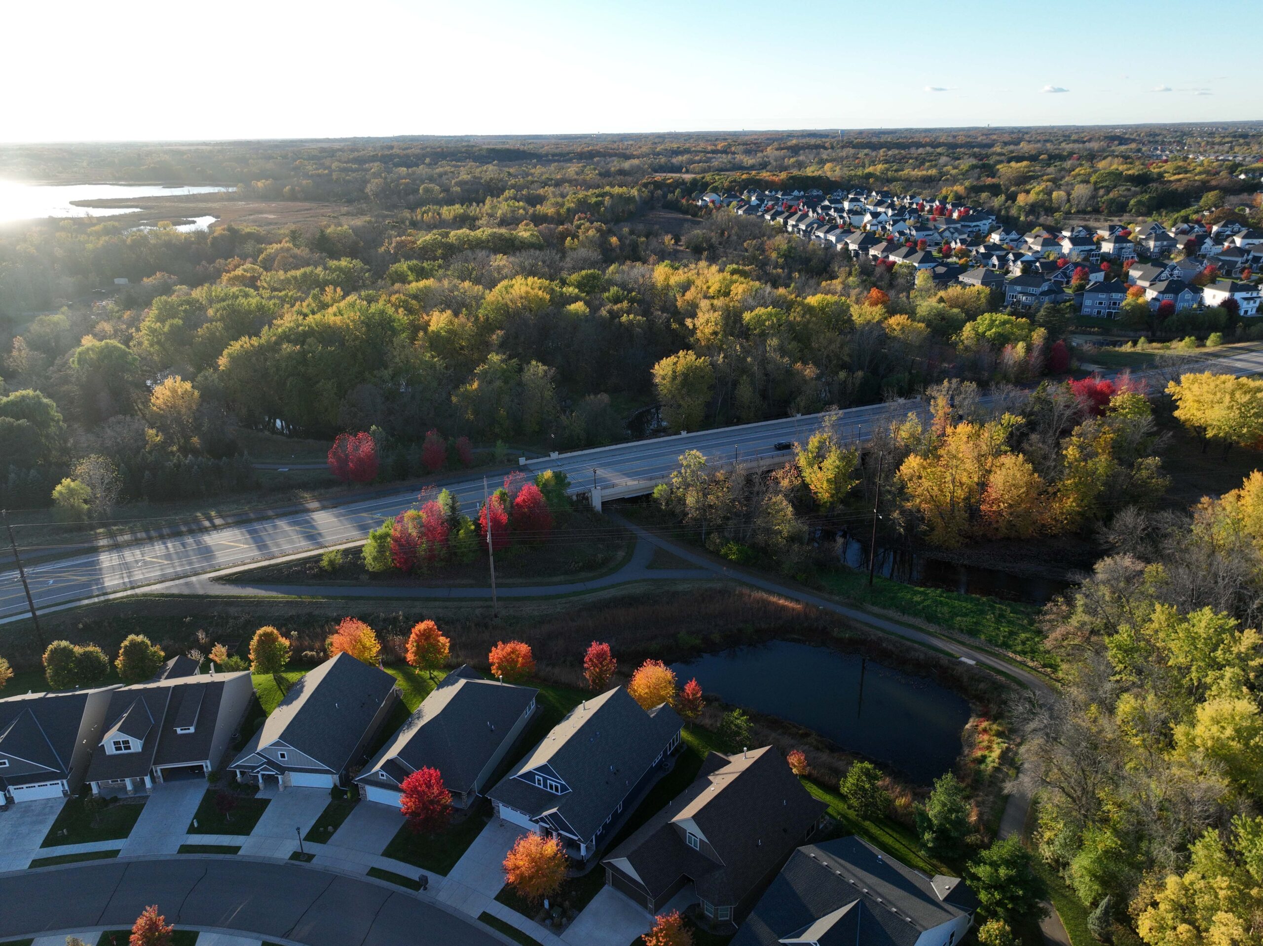 Aerial view of a residential neighborhood adjacent to a wooded area, roadway, and small pond with fall foliage.