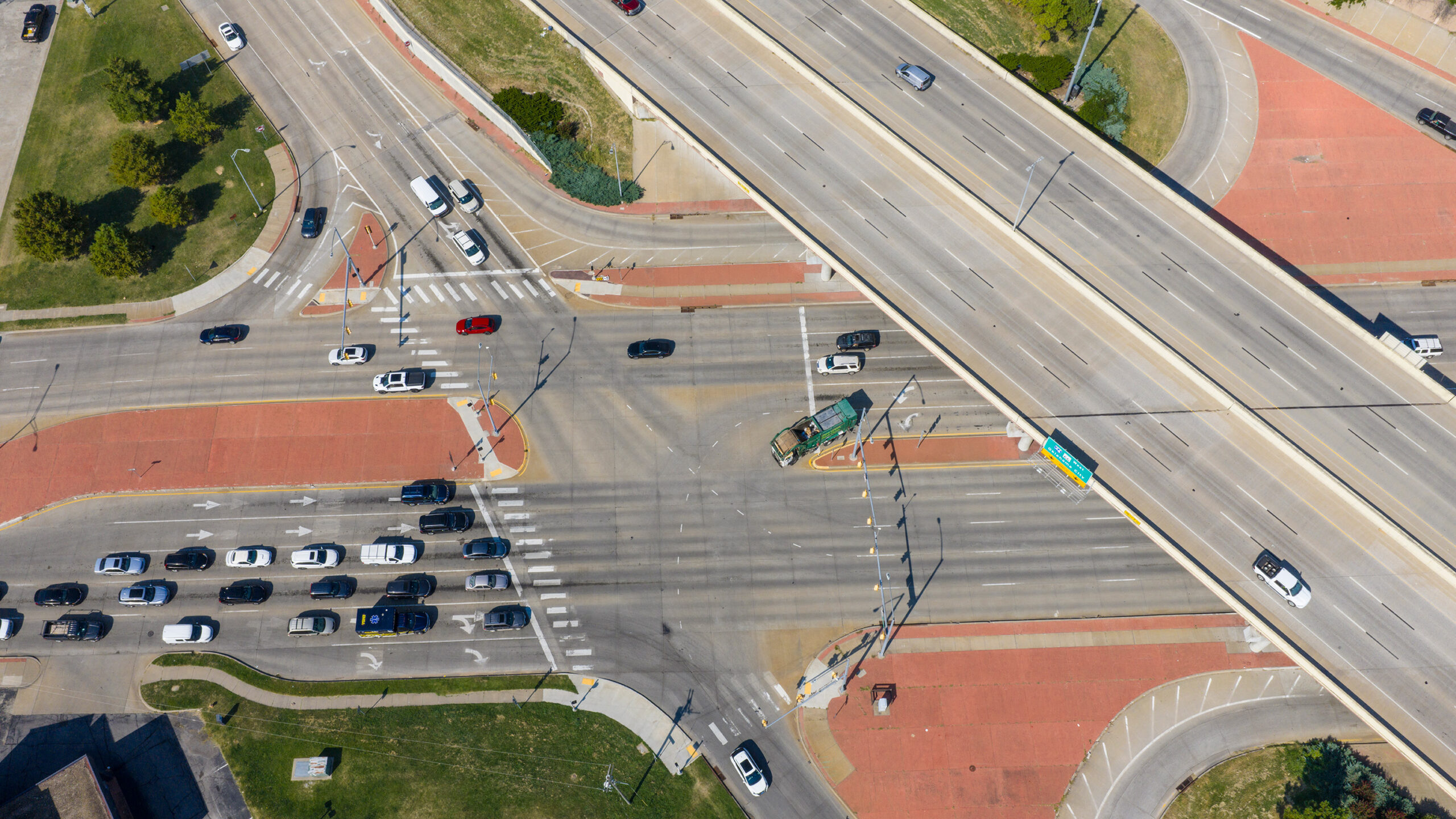 Aerial view of a large signalized intersection beneath a highway overpass, showing multiple lanes of vehicle traffic, crosswalks, turning lanes and landscaped medians.