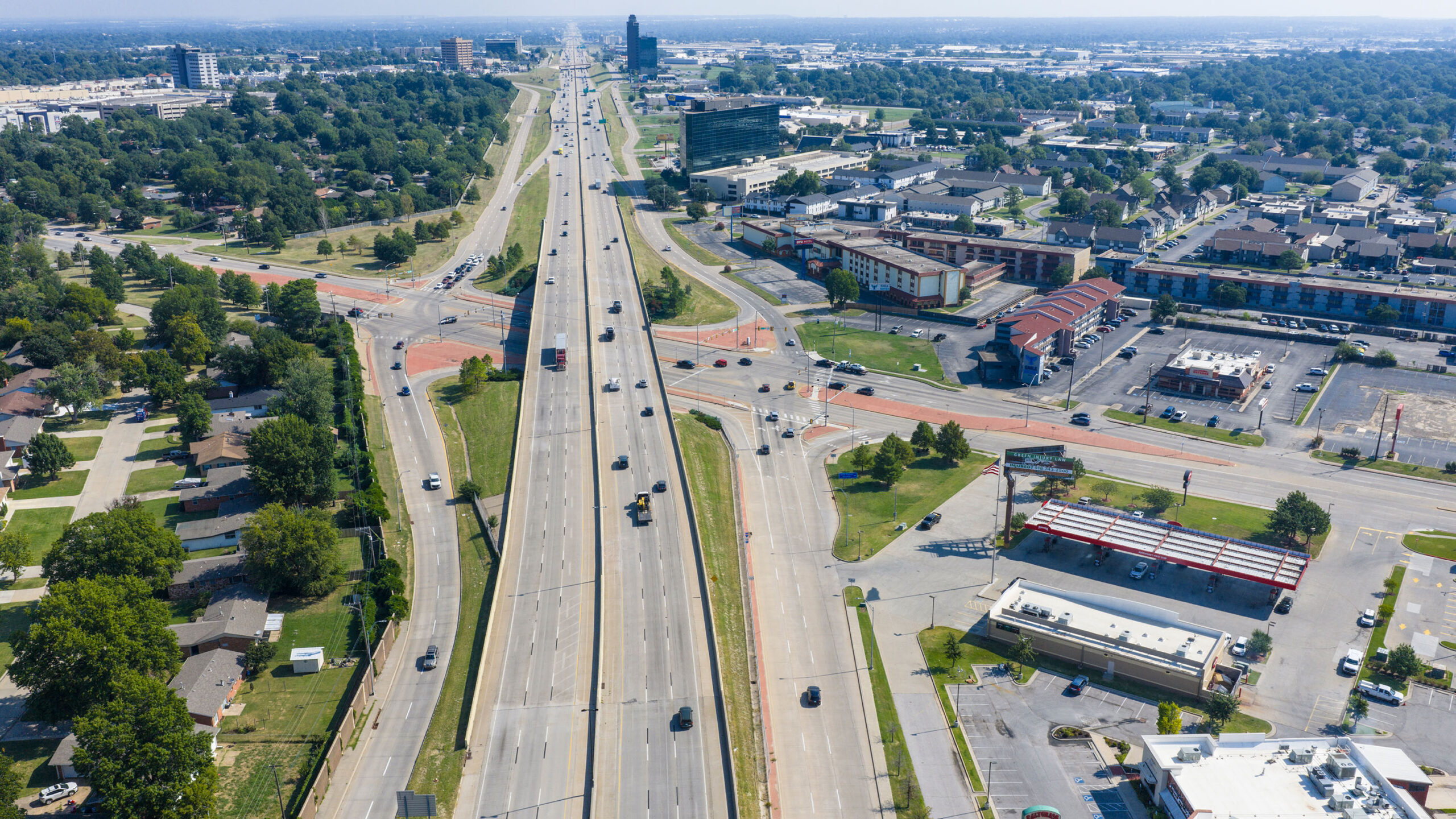 Aerial view of a multi‑lane divided roadway with a major intersection and highway overpass, showing vehicle traffic, turning lanes and surrounding commercial and residential areas.
