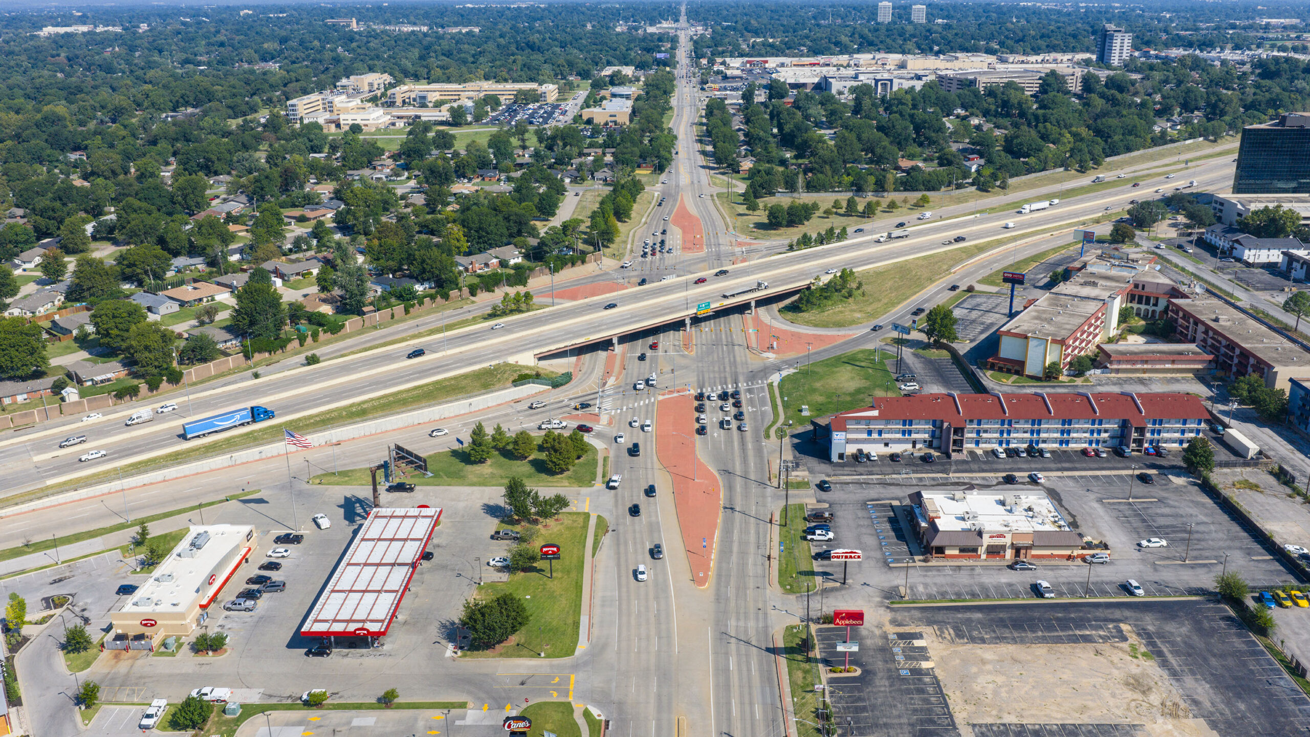 Aerial view of a multi‑lane roadway and highway overpass with vehicles moving through a large signalized intersection, surrounded by commercial buildings and residential neighborhoods.