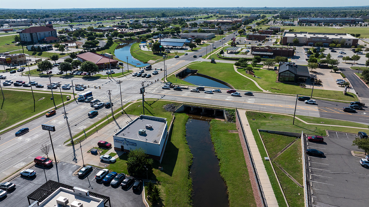 Aerial view of a roadway crossing over a concrete‑lined drainage channel, with surrounding commercial buildings, parking lots, sidewalks, and traffic lanes.